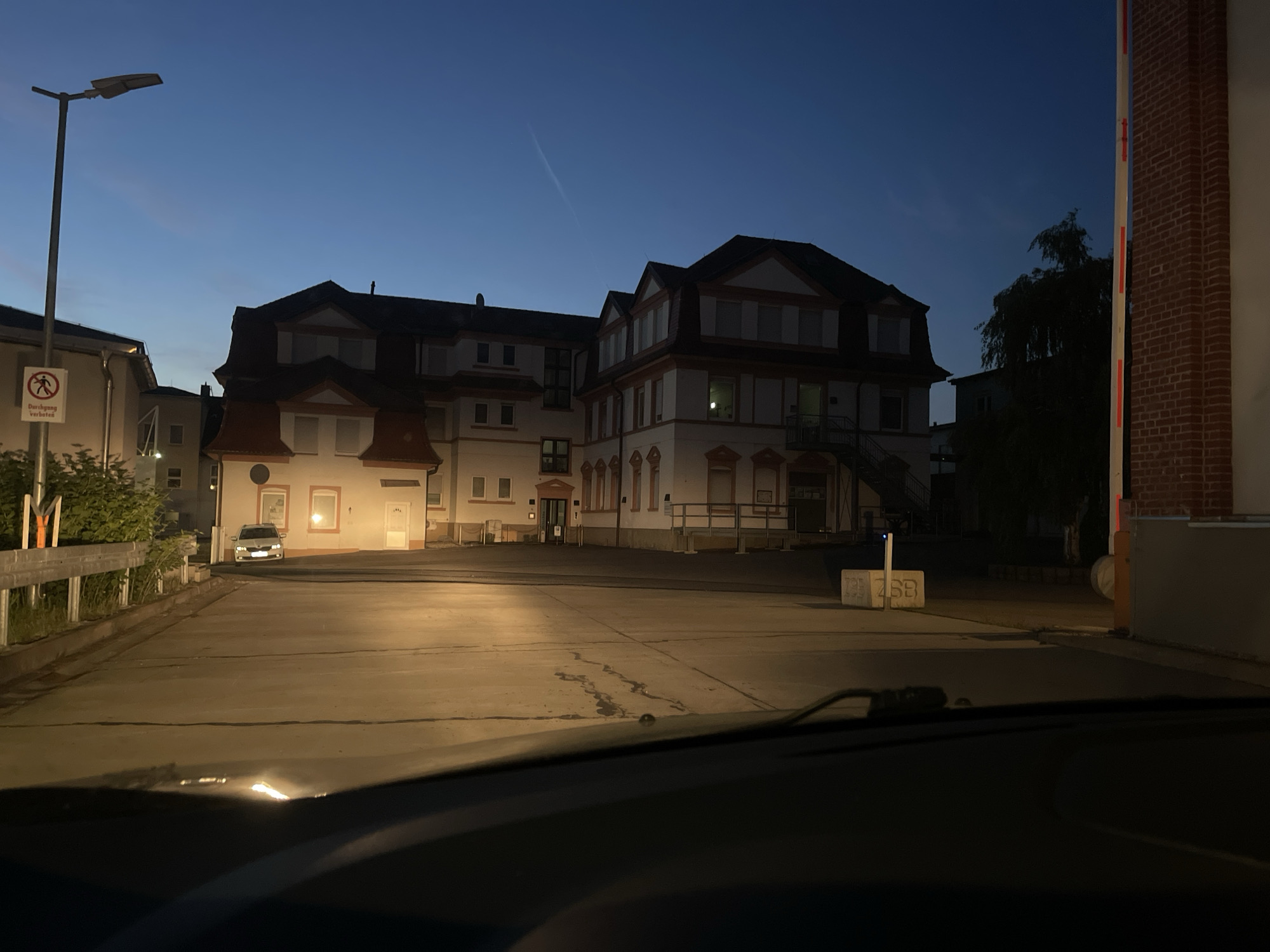 The image depicts a view from inside a vehicle, possibly a car, at night. The foreground is dominated by the dashboard and windshield of the vehicle. Through the windshield, a concrete parking area stretches towards a large, three-story building with white walls and dark roofs. A streetlight stands to the left of the building. A pedestrian crossing sign is visible near the streetlight. A vehicle is parked near the left side of the building. The background sky transitions from a lighter hue near the horizon to a darker blue above. A portion of a brick and concrete structure is visible on the right. The location appears to be Unknown, Unknown.