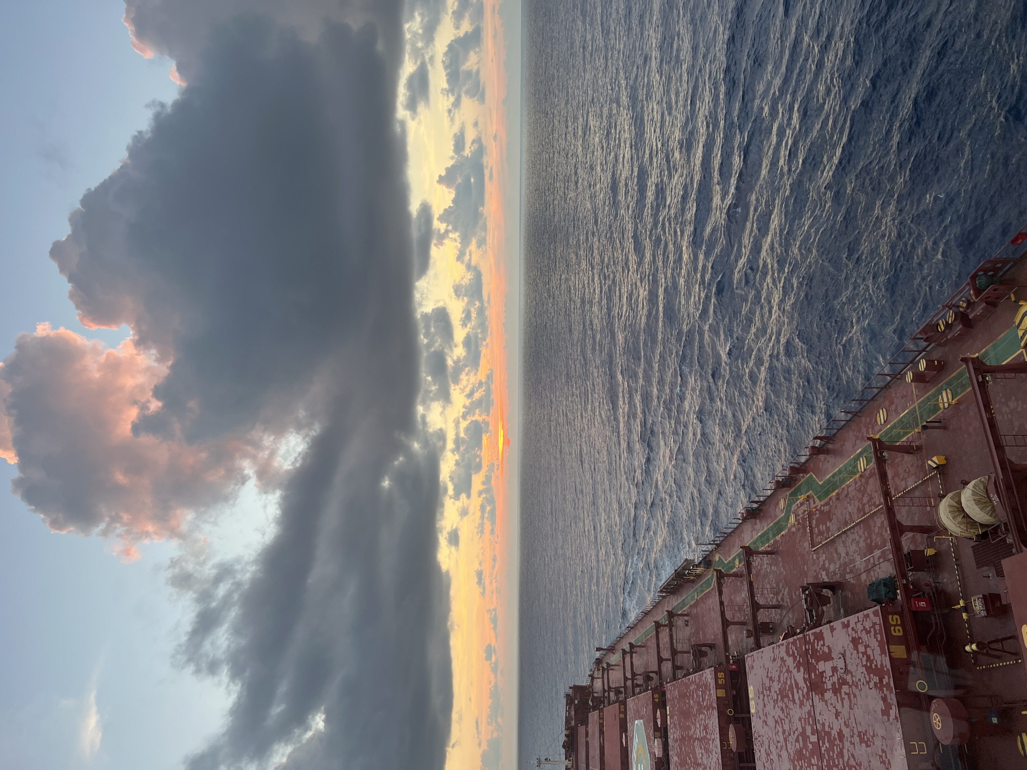 A high-angle medium shot captures the deck of a large cargo ship at sea, with the ocean and a cloudy sunset filling the upper half of the frame. The ship's deck is a deep red, with visible hatches, railings, and various equipment. The ocean is a deep blue, with small waves and whitecaps. The sky is a blend of pale blue, orange, and pink, with large, dark gray clouds dominating the upper portion of the image. The sun is a small, bright orange disc peeking through a gap in the clouds near the horizon. The scene appears to be set at dusk or dawn.