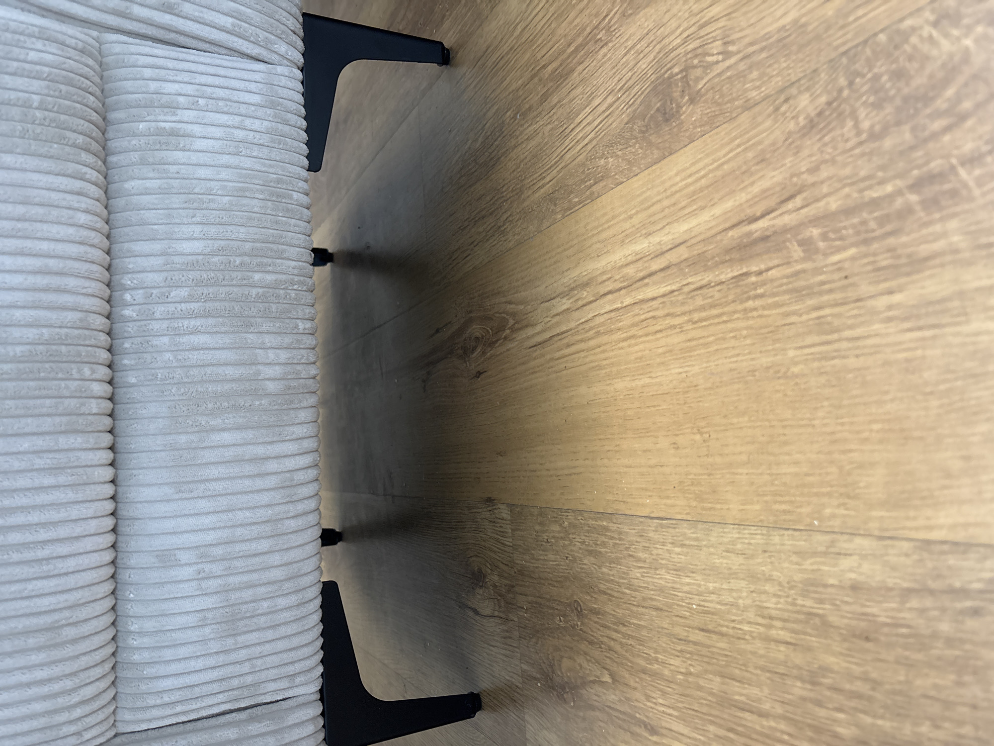 The image shows a close-up, low-angle view of the legs of a light-colored, corduroy upholstered piece of furniture, possibly a sofa or chair, resting on a light brown wooden floor. The furniture legs are black and have a simple, modern design, with two legs visible on the left side of the frame and one on the right. The corduroy texture of the furniture is prominent, with distinct horizontal ridges running across its surface. The wooden floor has a natural grain pattern and appears to be made of planks. A shadow is cast by the furniture onto the floor, suggesting the presence of overhead lighting. The overall scene is static and focuses on the intersection of the furniture and the floor. There is no text visible in the image.