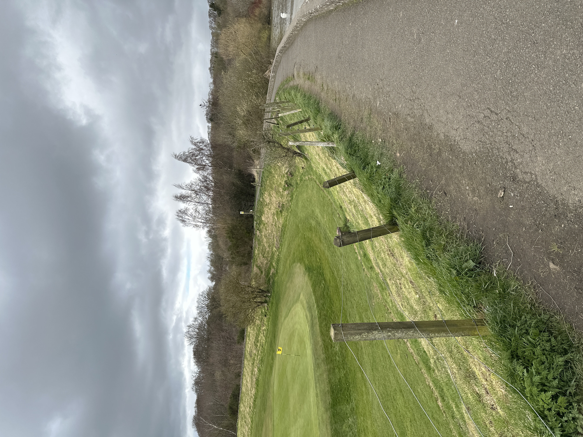 The image depicts a golf course on an overcast day. A path runs along the right side of the frame, separated from the fairway by a wooden post and wire fence. The fairway is green and well-manicured, with a yellow flag indicating the location of the hole. Trees line the left side of the frame. The sky is filled with gray clouds. The image was taken in Dunfermline, United Kingdom.