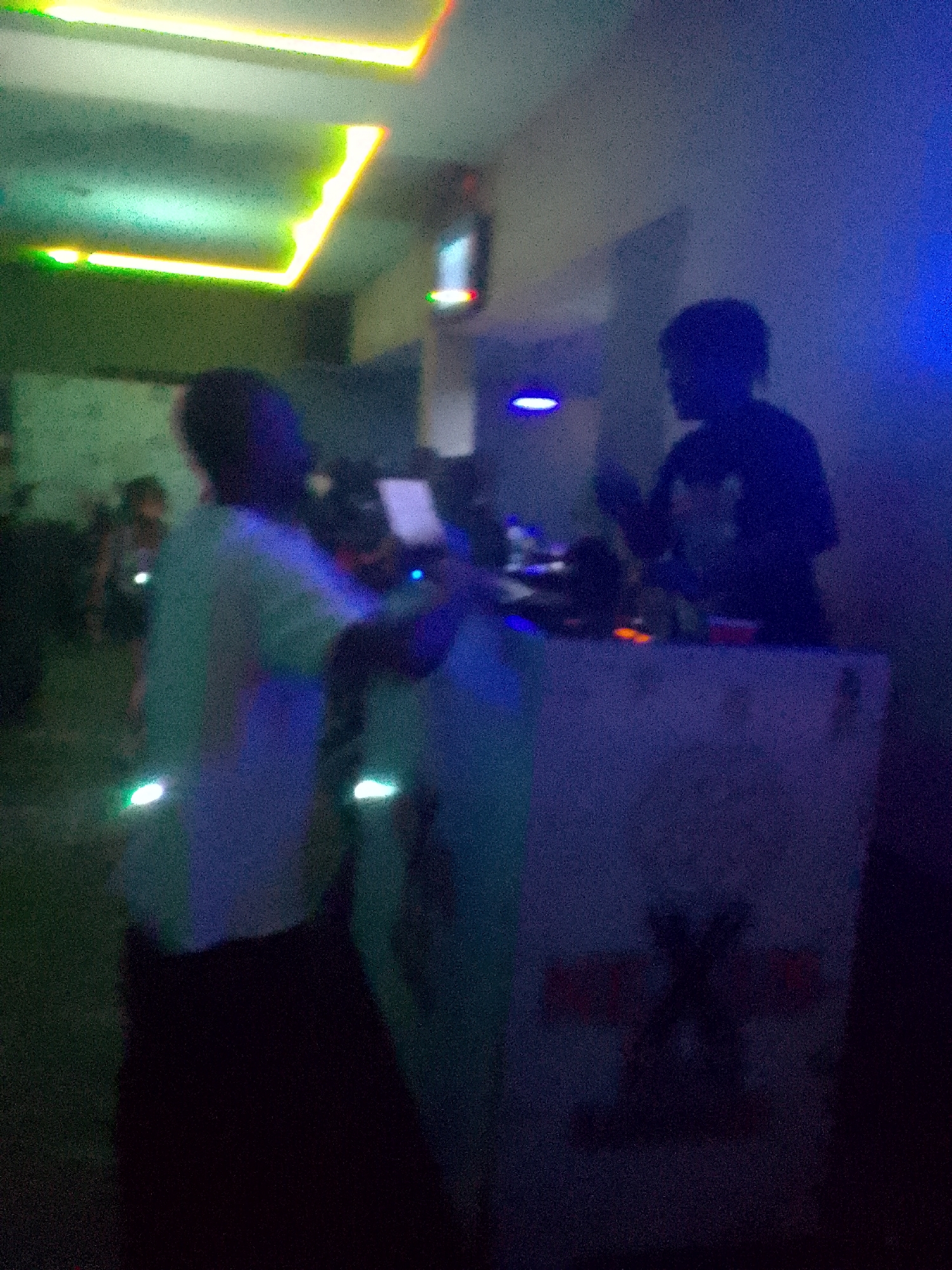 The image captures a blurry, low-light indoor scene, characteristic of a nightclub or bar environment. Several individuals are present, mostly appearing as indistinct silhouettes or blurred forms due to the poor lighting and image quality.
In the foreground-middle, a person wearing a light-colored shirt is seen from behind, facing away from the camera. This individual has two prominent, bright, possibly reflective or glowing dots on their lower back. To the right, another person, likely a DJ, is silhouetted behind a counter or DJ booth, appearing to be engaged with equipment. Other blurred figures are visible in the background, suggesting a crowd.
The setting is dark, primarily illuminated by artificial lights. Green and yellow neon strip lights are visible on the ceiling, casting a glow. Blue ambient lighting is present on the right side of the frame, and several small, bright light sources are scattered throughout. The overall activity appears to be a social gathering or a musical performance, with a DJ at the center.
The image is significantly grainy and out of focus, indicating low-light conditions and possibly camera movement during capture. On the front of the DJ booth, there are faint, dark markings or a stylized graphic, but no clear, readable text is discernible. The time of day is clearly night.