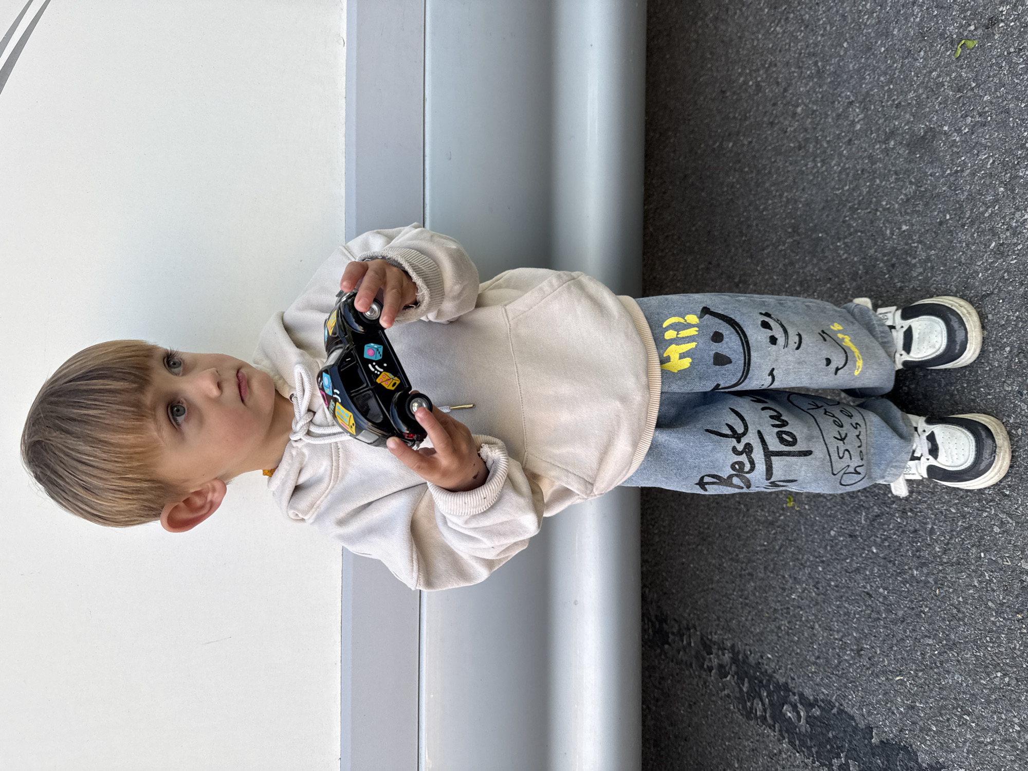 A young child with blonde hair, wearing a light beige hoodie and jeans with cartoonish graphics (smilies and the words 'Best Town' and 'Ski House' printed on them) and black and white sneakers, is standing on an asphalt surface, next to a white and grey metallic panel, possibly a wall. The child is holding a black toy car in their hands. The location is unknown; there is no contextual information regarding the city of Riedlingen, Germany.