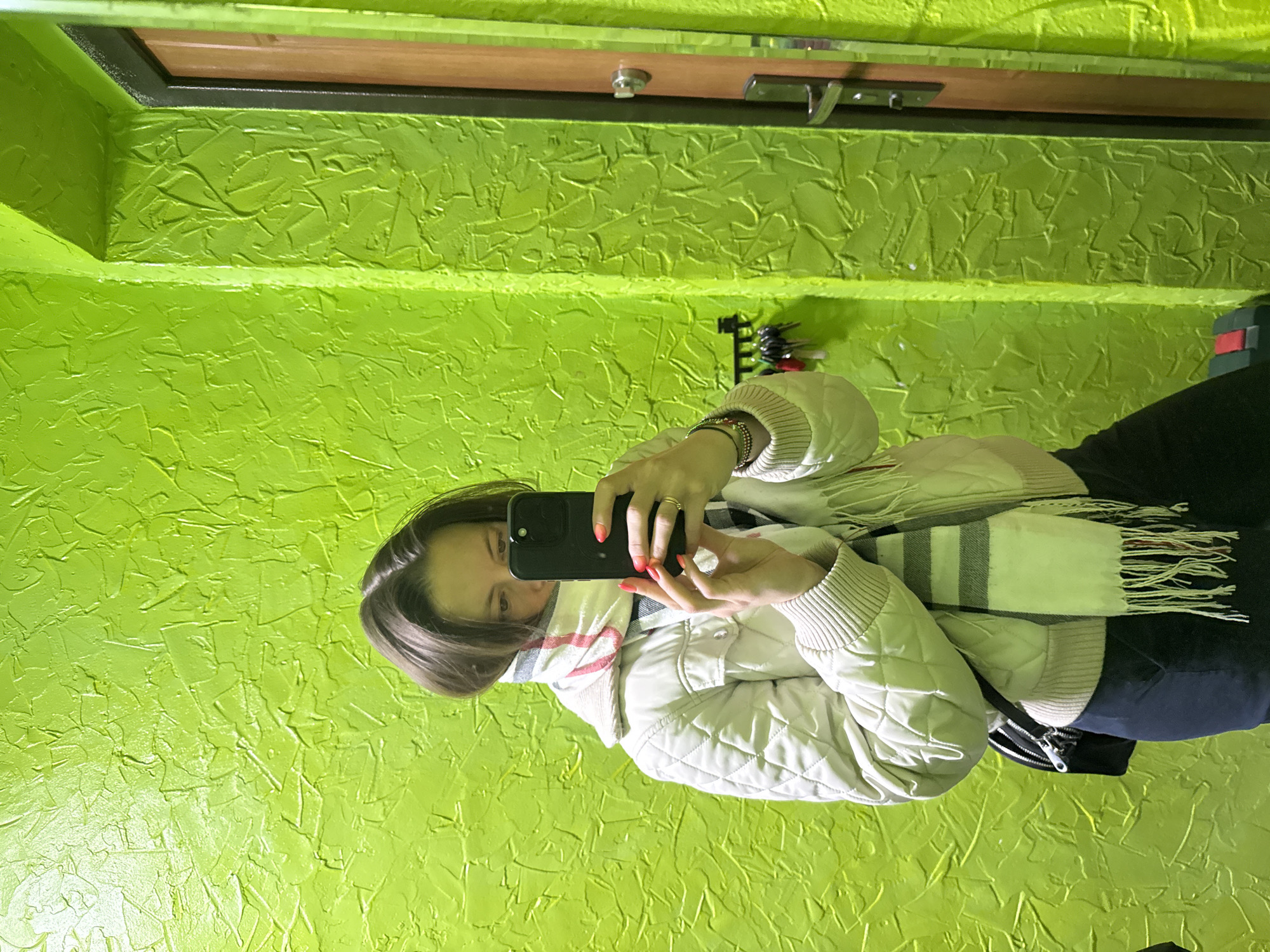 This image captures an indoor mirror selfie taken by a young female. She is seen holding a black smartphone with both hands, its screen reflecting her face, which is partially obscured by the device. She has dark hair, and her fingernails feature an ombre polish, transitioning from red to orange. On her right hand, a ring is visible, and her left wrist is adorned with bracelets.
She is dressed in warm attire, including a light-colored, possibly beige or off-white, quilted jacket with ribbed cuffs, and a striped scarf with white, black, and reddish-pink bands, completed with tassels. She appears to be wearing dark pants.
The setting is an interior space, likely a hallway or entryway, distinguished by vibrant, almost neon, lime green walls with a pronounced textured finish, resembling stucco. In the background, seen in the mirror's reflection, there is a wooden door with a metallic handle and a lock, framed by a dark border. To the right, a small rack holding several keys is also visible. The lighting is bright and even, consistent with artificial indoor illumination. The warm clothing suggests cooler weather conditions. No text is visible in the image. The provided location context is Chaikovsky, Russia.