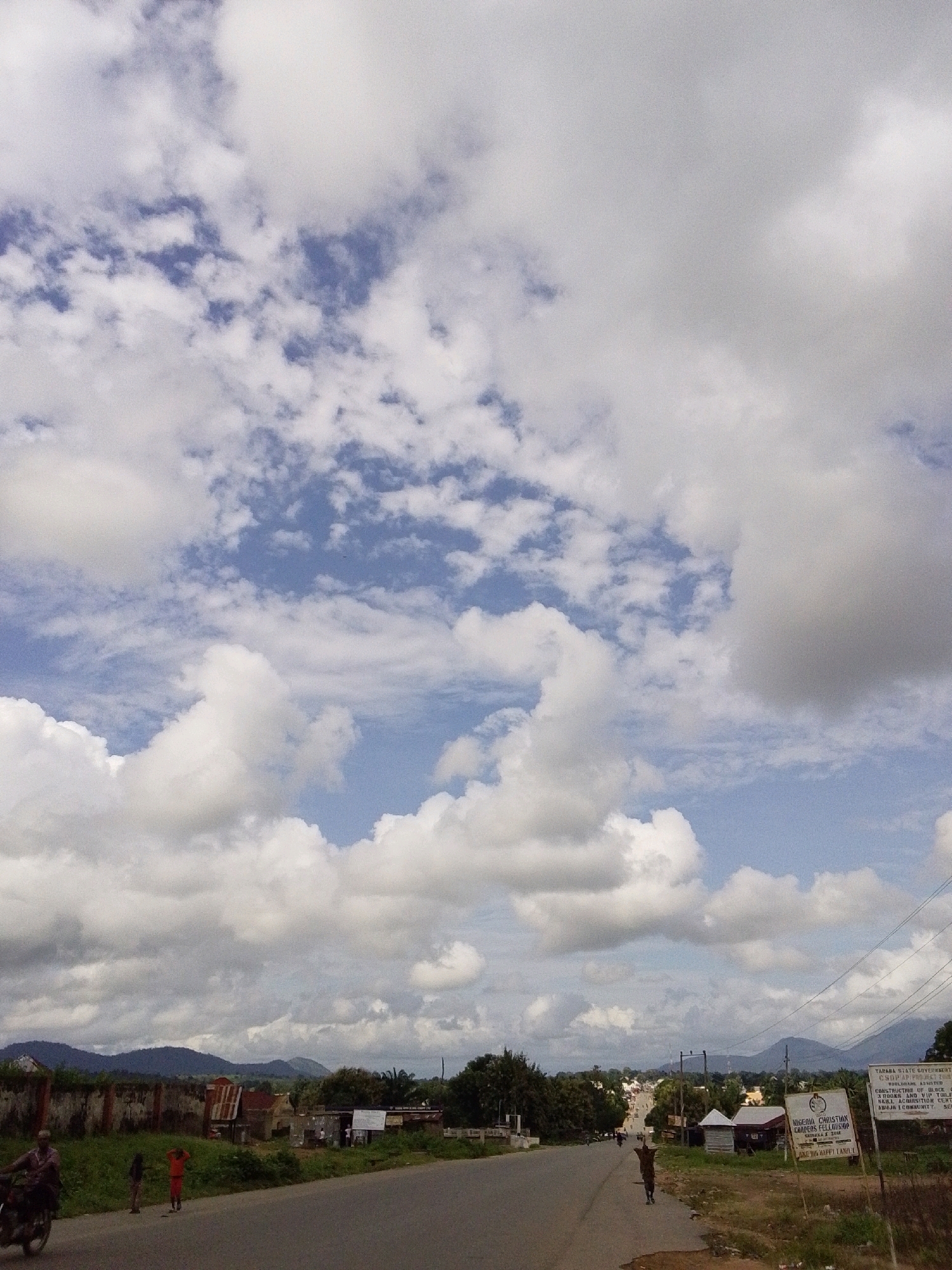 This image depicts a street scene in Serti, Nigeria, under a partly cloudy sky. The road occupies the foreground, with the sky filled with dramatic white clouds against patches of blue. On the left side of the road, a motorcycle is partially visible with a rider. Further down the road, two children are standing, one in an orange shirt and the other in dark clothing. A low wall runs along the left side of the road, with some buildings and structures visible behind it, including a building with a corrugated metal roof. Trees and vegetation are present along the side of the road. On the right side of the road, there are two signs. One sign, white with black text, appears to be an official notice, possibly related to government projects or initiatives in the Taraba State. The second sign, also white with black lettering, reads "NIGERIA CHRISTIAN CAMPERS FELLOWSHIP" and has additional text below. A person is walking away from the camera on the right side of the road. In the distance, a town is visible with more buildings and trees, and beyond that, rolling hills and mountains form the horizon. The overall impression is a rural or semi-urban setting on a bright, possibly warm day.