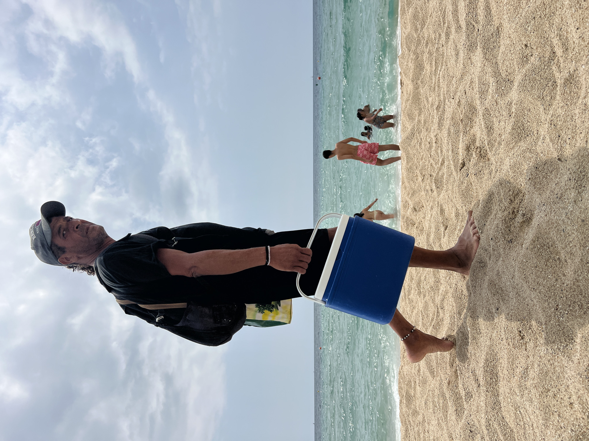 The image depicts a man walking on a sandy beach in El Masnou, Spain, carrying a blue cooler and a backpack. He is wearing a black t-shirt, black shorts, and a gray baseball cap. His right foot is raised as he walks towards the camera. In the background, several people are in the turquoise ocean, some wading and others swimming. The sky is partly cloudy.