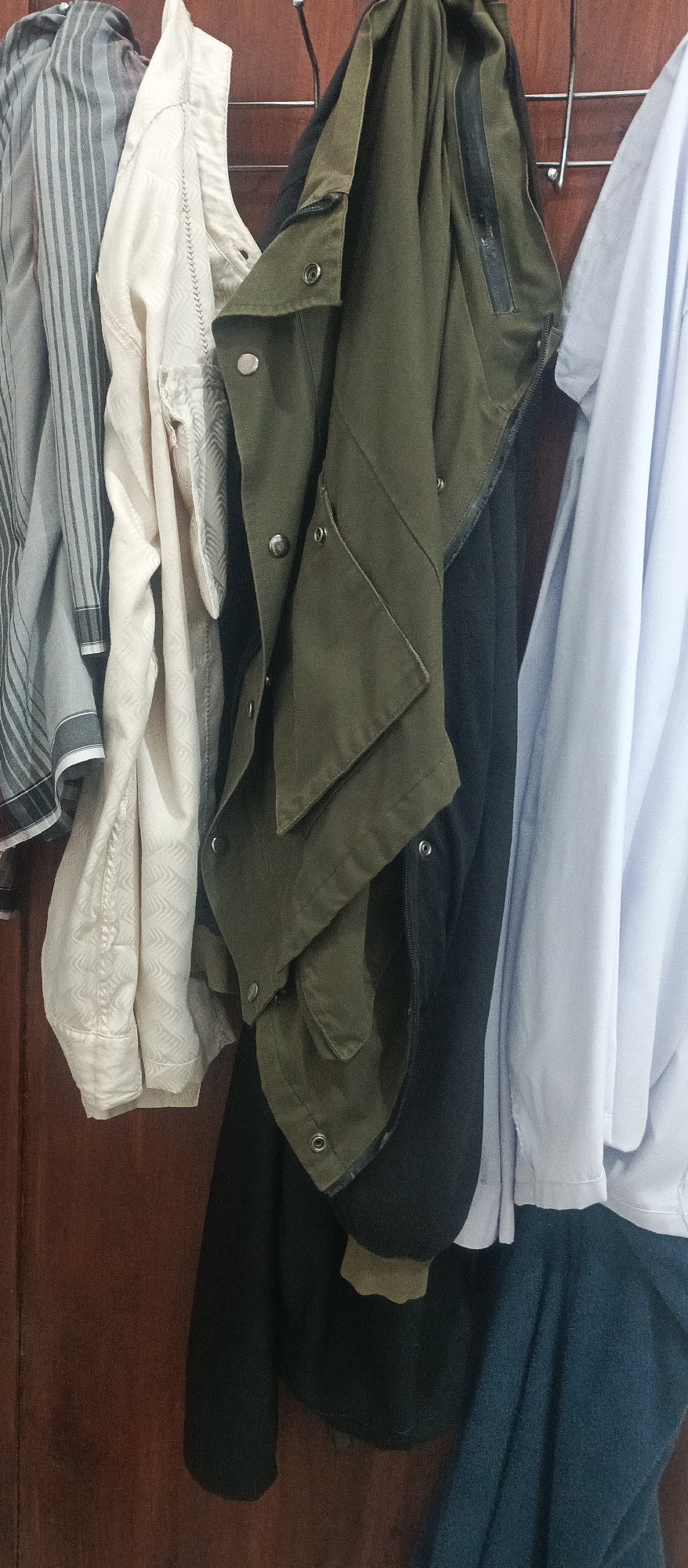 This is a close-up, waist-up, full-body shot of several pieces of clothing hanging on a rack against a wooden wall, likely in Arjasa, Indonesia. From left to right, there is a gray and white striped shirt, a light cream-colored shirt with a subtle patterned texture, a dark olive green jacket with metal snap closures, a black garment, and a light blue long-sleeved shirt. The olive green jacket is draped open, revealing some of the black garment beneath. The clothing appears to be casually hung, suggesting a domestic or casual setting. The background is a plain wooden surface.