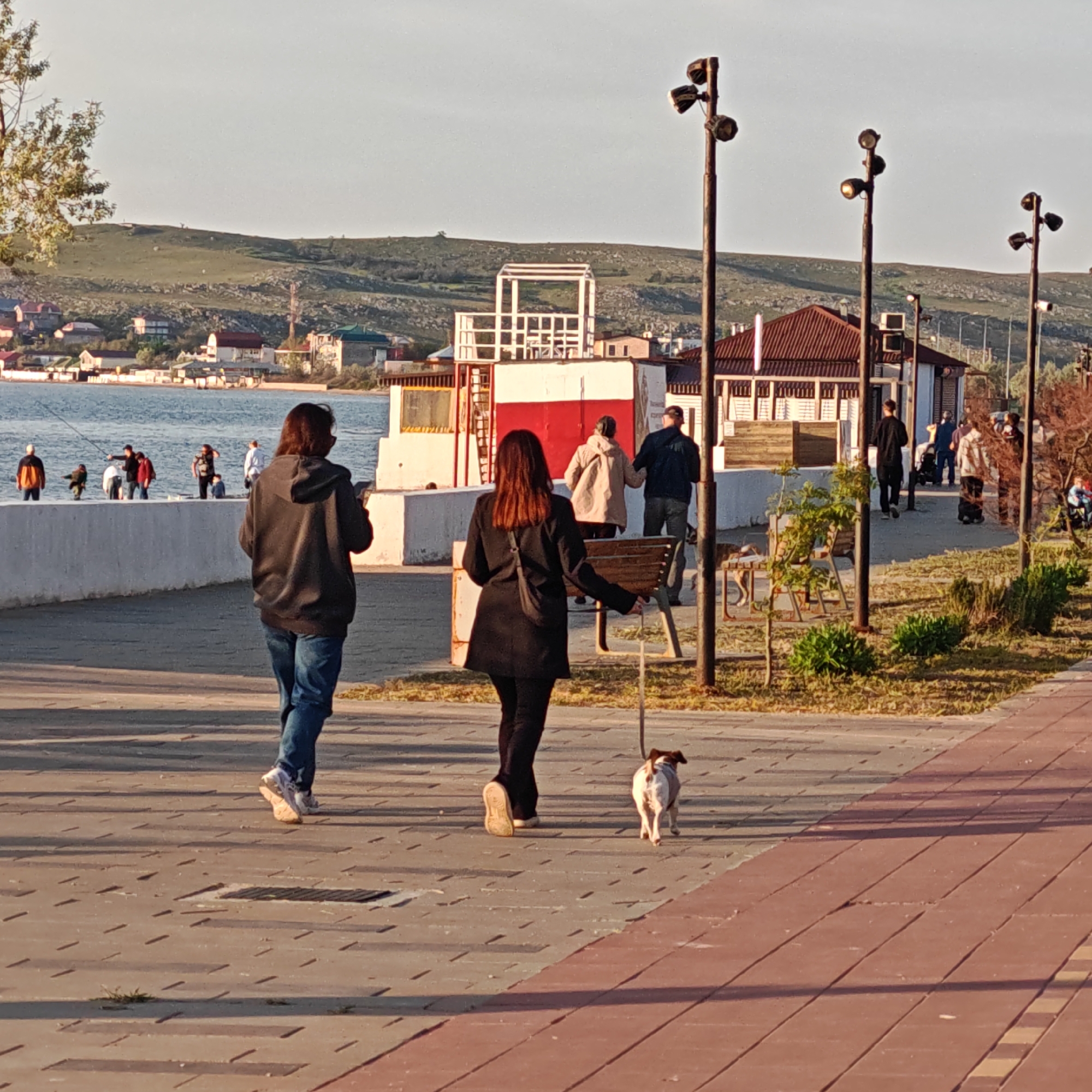 The image captures a scene from городское поселение Щелкино, Ukraine. It depicts a walkway made of interlocking pavement tiles with some vegetation and trees visible on the right. A man and a woman are walking away from the viewer on the walkway. The woman has a dog on a leash. In the background is a white building with red trim, streetlights, and a slight rise in the terrain with houses and a body of water visible in the distance. People are gathered near the water's edge and some are fishing.