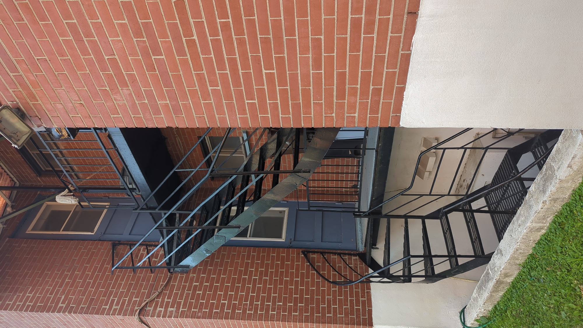 This is a photograph of the exterior of a brick building. A black metal fire escape staircase is attached to the side of the building. The staircase is a combination of straight sections and a spiral staircase. A blue door with a window is visible on the second level of the building, accessed by the fire escape. A mop head is hanging on the railing of the fire escape. The building is made of red brick, and the lower portion of the wall is painted white. There is some green grass and a concrete ledge in the foreground. The photograph appears to have been taken during daylight hours. There is no information available about the specific location in the city of Montreal, Canada, or any people or actions.