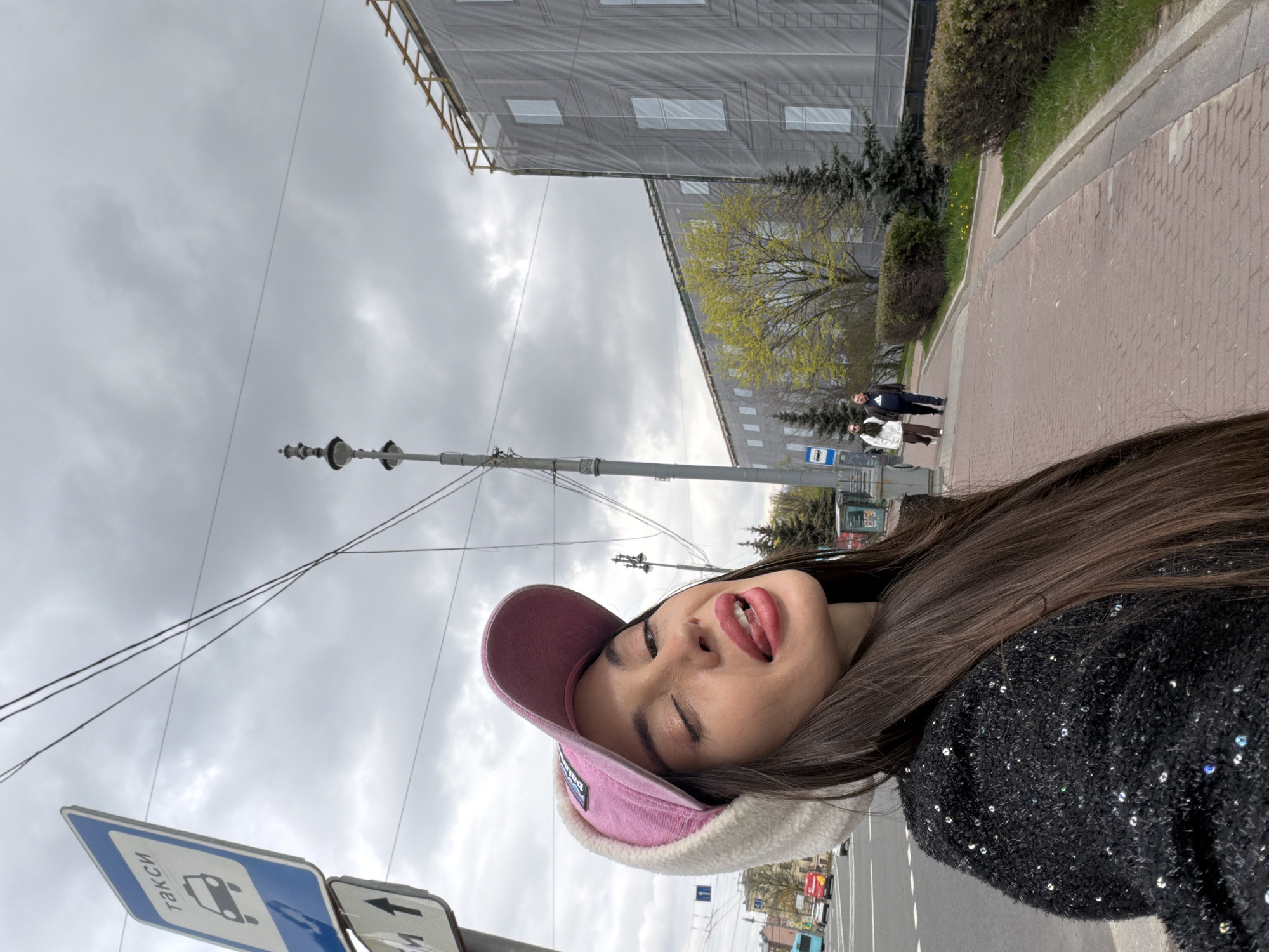 The image shows a young woman with long brown hair wearing a pink cap and a black sequined jacket. She is standing on a sidewalk with a building under construction in the background. There is a street sign visible that is in Russian. A man and woman can be seen in the distance. The location is Saint Petersburg, Russia.