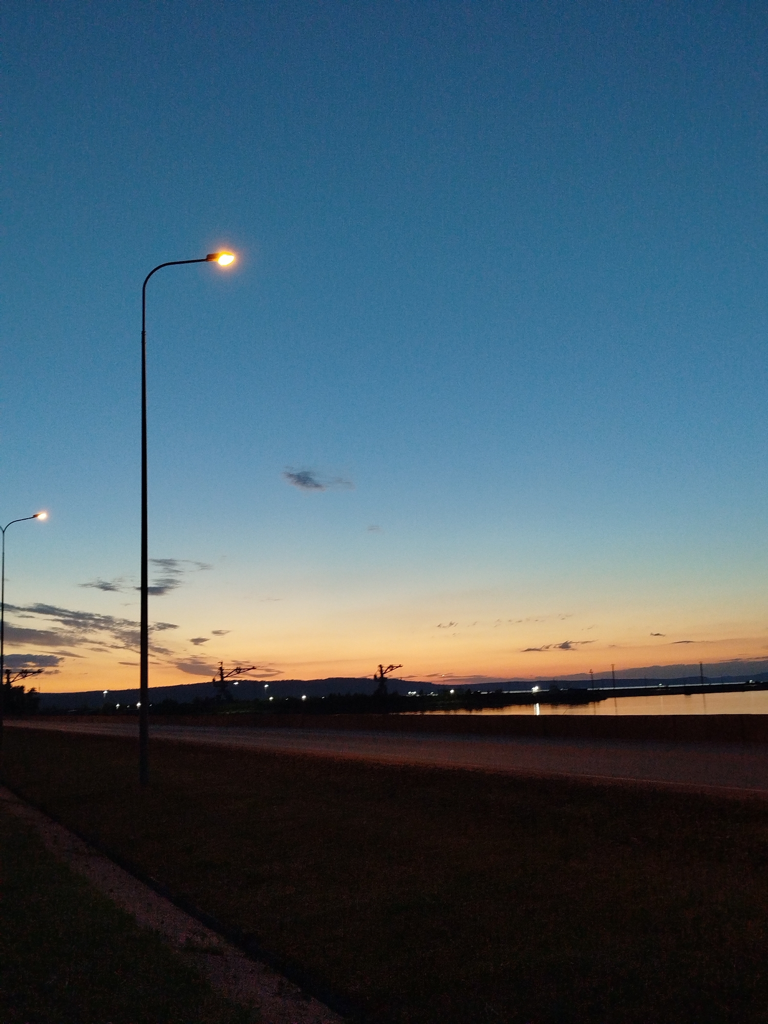 The image depicts an outdoor scene at dusk, with a clear gradient in the sky transitioning from a deep blue at the top to a warm orange and yellow near the horizon. Several streetlights are visible, one prominently in the left foreground, emitting a bright yellow light. Another streetlight is partially visible further to the left. Silhouetted against the horizon are distant hills or mountains, and a body of water, possibly a lake or the sea, reflects the ambient light. There are also some man-made structures, possibly power lines or towers, visible in the distance. The foreground is dominated by dark grass and a paved area, possibly a road or sidewalk. The overall impression is a tranquil, transitional moment between day and night in an unspecified urban or semi-urban setting.