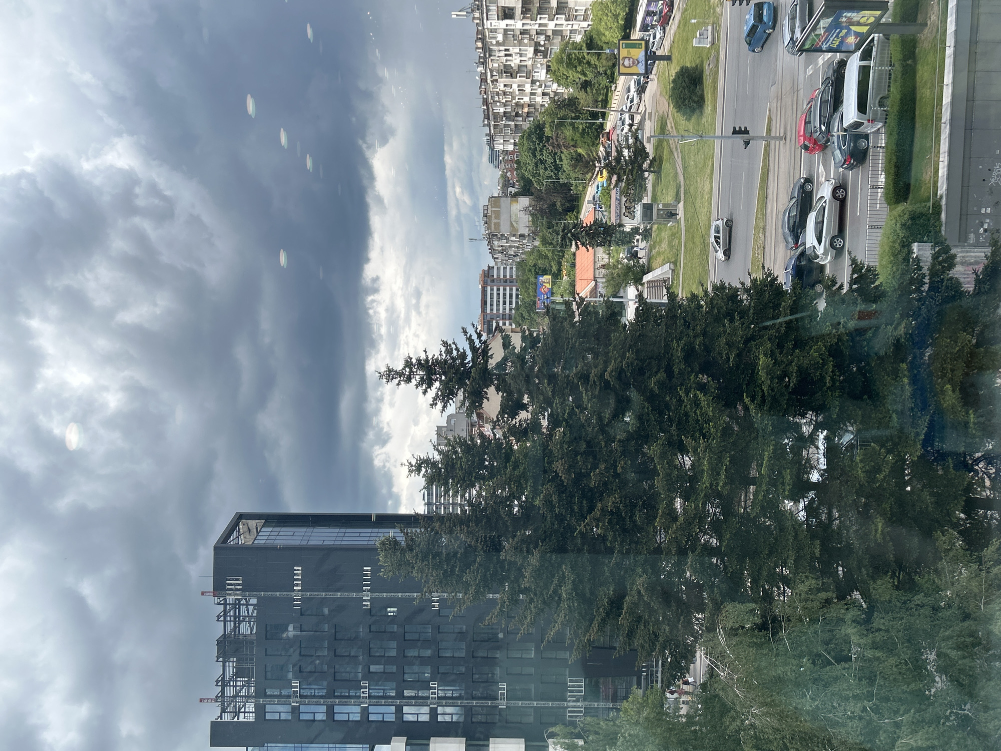 The image depicts a cityscape under a cloudy sky. A modern black building stands prominently in the foreground, partially obscured by tall green trees. Beyond the trees and the building, a road with several cars and green grassy areas runs parallel to a row of older, high-rise apartment buildings. Traffic lights are visible on the road. The location is Unknown, Unknown. The overall impression is of a mix of urban development and natural elements.
