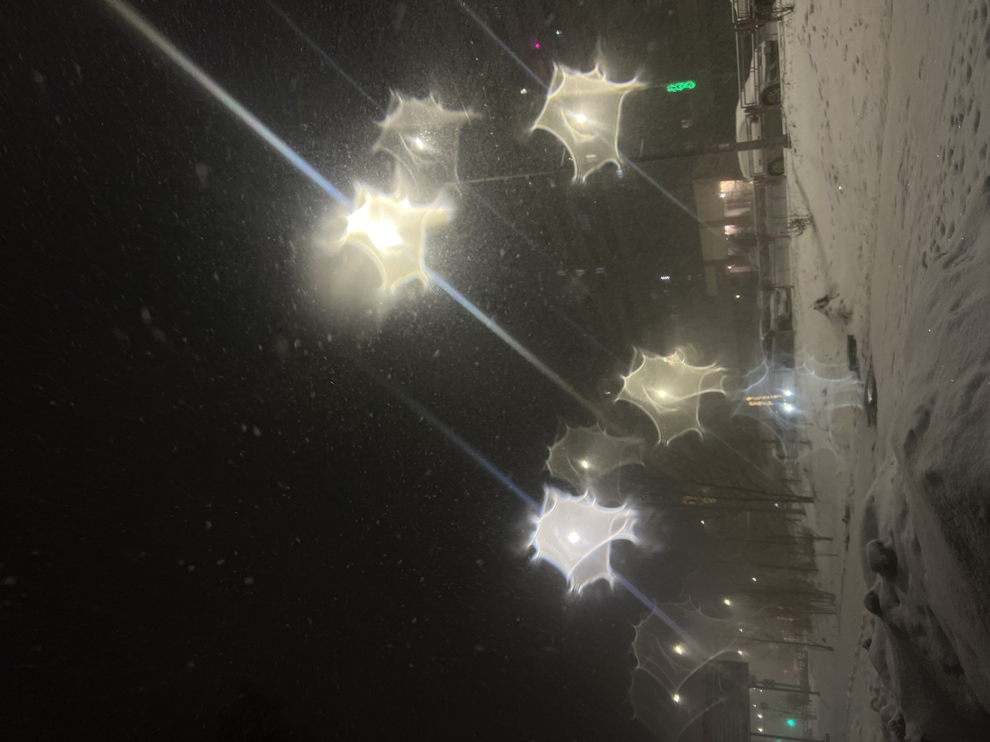 The image depicts a nighttime scene in what appears to be a snowy urban environment. The view is partially obscured by a dirty or wet surface, possibly a window, which creates star-shaped reflections or refractions of the lights in the background.
On the right side of the frame, there is a snow-covered area, likely a roadside or sidewalk. A vehicle is parked in the snow. In the background, a building with illuminated windows can be seen. The overall impression is one of a winter night in a town or city. There is a hint of green light and other building lights.
Based on the provided information about the location being Ust-Ilimsk, Russia, it can be assumed that the image captures a typical winter scene in that city.