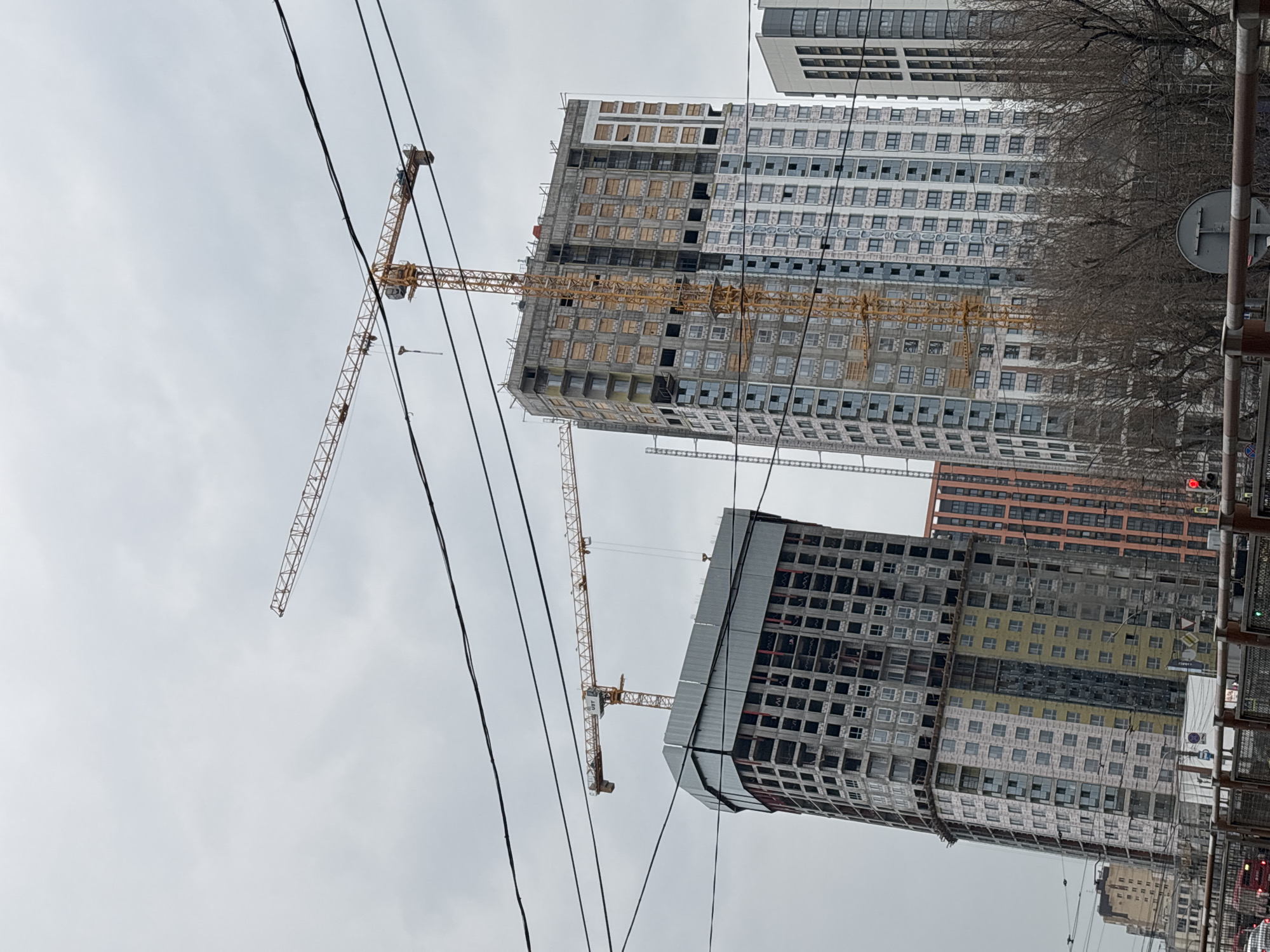 The image captures an urban construction site in Perm, Russia. Several high-rise buildings under construction dominate the view, with their unfinished facades partially covered or boarded up. Yellow construction cranes are strategically positioned around the buildings, actively participating in the ongoing work. Power lines crisscross the foreground. The sky is overcast, and a few bare branches of trees are visible, suggesting either late autumn or early spring. The presence of traffic lights and street signs hint at an active street or intersection in the foreground.