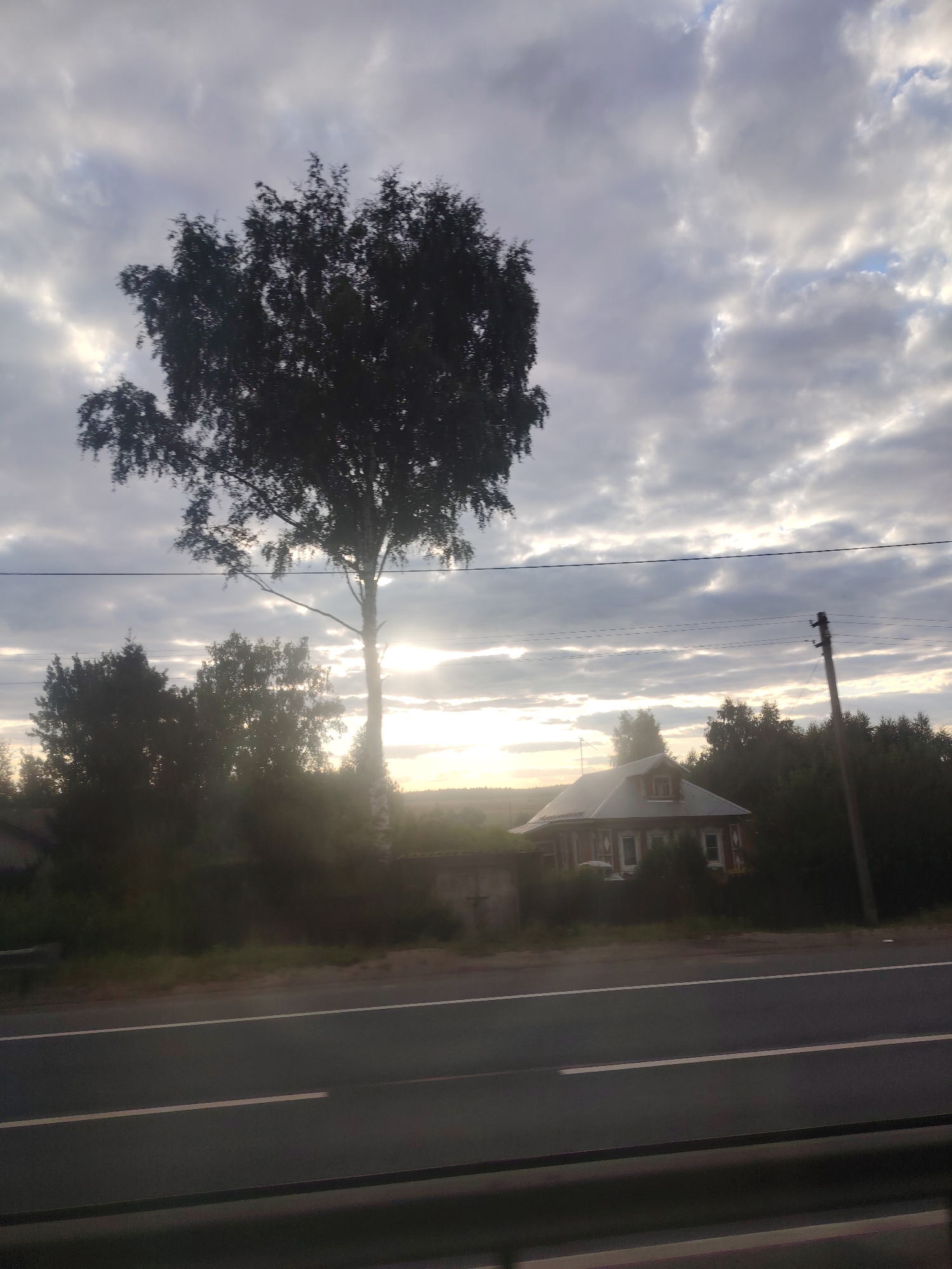 The image depicts a rural scene with a road in the foreground. A tall, leafy tree stands prominently on the left side of the frame, silhouetted against a cloudy sky with the sun breaking through. In the middle ground, a rustic-style house with a red roof and white trim is visible, surrounded by greenery. A utility pole with wires runs across the scene. The lighting suggests it is either sunrise or sunset, with a soft glow illuminating the sky and casting shadows. The location is Zavidovo, Russia.