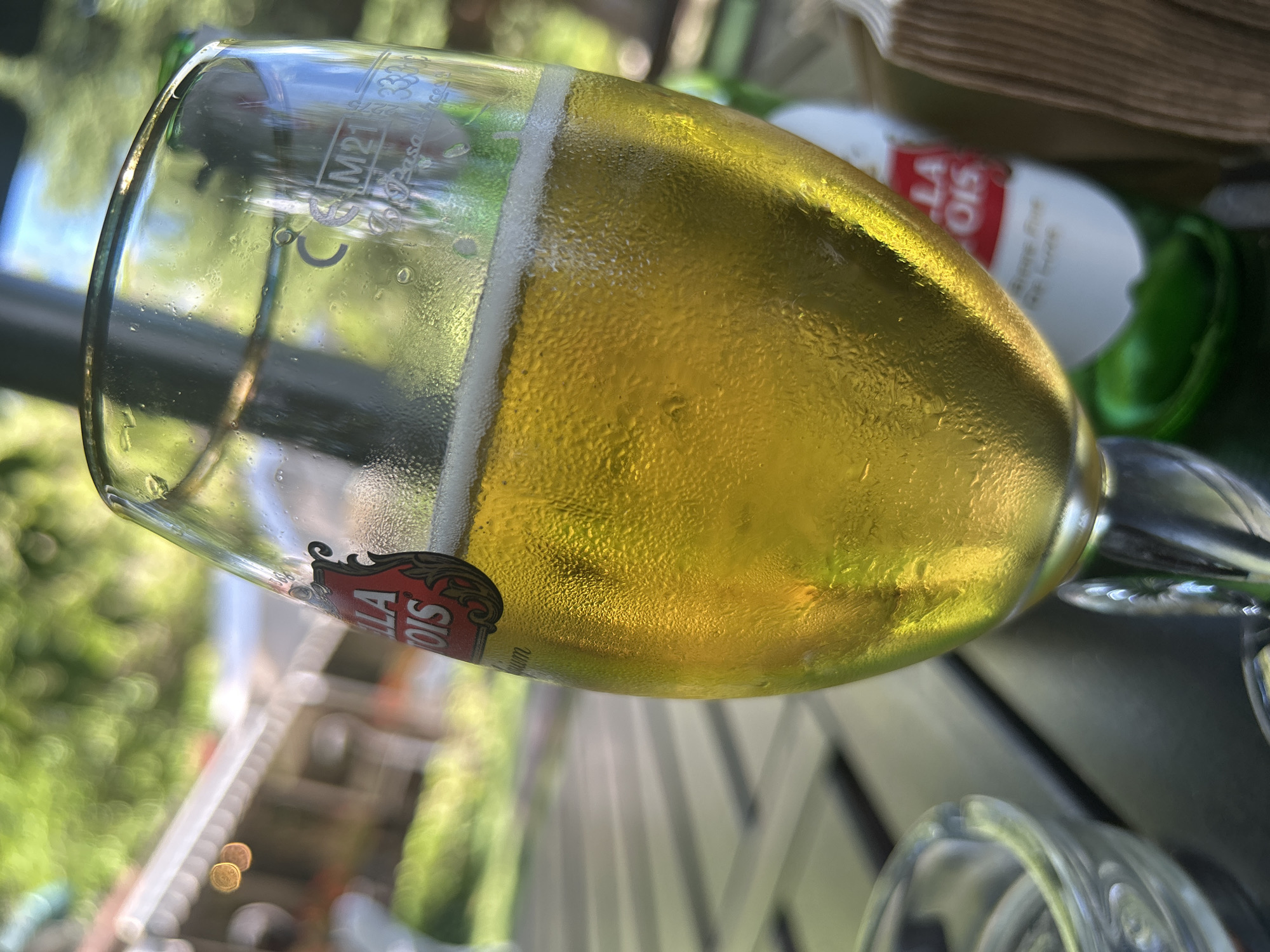 The image shows a close-up of a glass of Stella Artois beer on a dark, slatted table. The beer is golden yellow and appears to be cold, as evidenced by condensation on the outside of the glass. A red Stella Artois label is visible on the glass. In the background, out of focus, are lush green trees and a building with a light-colored roof. A bottle of Stella Artois is partially visible behind the glass. The scene suggests an outdoor setting, possibly a restaurant or cafe, during daylight hours. The location is identified as Varna, Bulgaria.