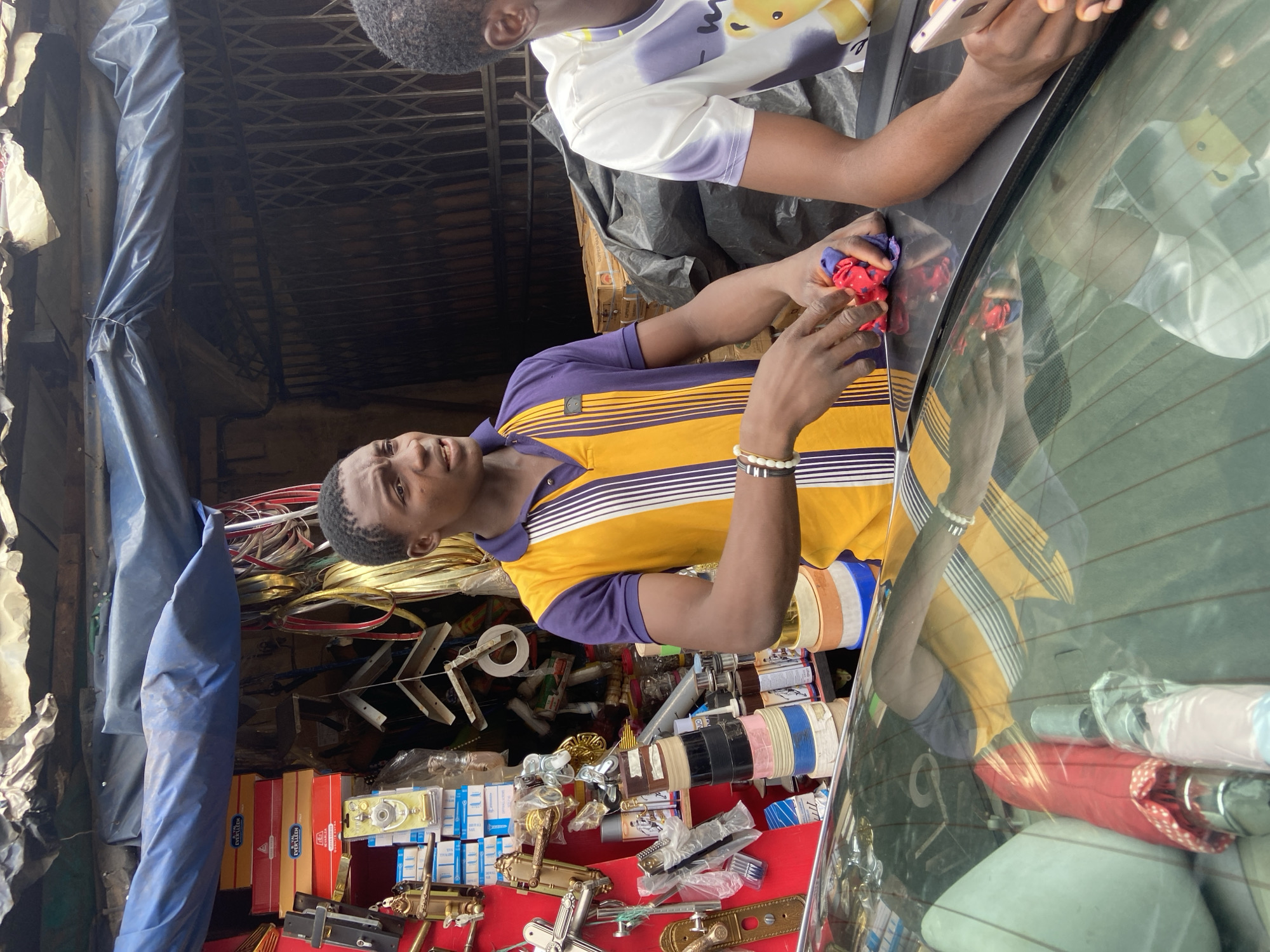 In Ogidi, Nigeria, a young man wearing a yellow and purple polo shirt stands in front of a market stall that sells various hardware items. The stall's shelves are stocked with door handles, locks, boxes, and rolls of tape. The man is holding a red and blue cloth in his hands, appearing to be folding or cleaning it. To his right, another young man, partially visible, is holding a smartphone. The scene suggests a commercial interaction or a moment of pause at a busy marketplace. The background shows a corrugated iron roof and a metal gate, typical of many shops in the area. The reflection on the car windshield in the foreground offers a distorted view of the surroundings, including more of the market stall's inventory and the people present.