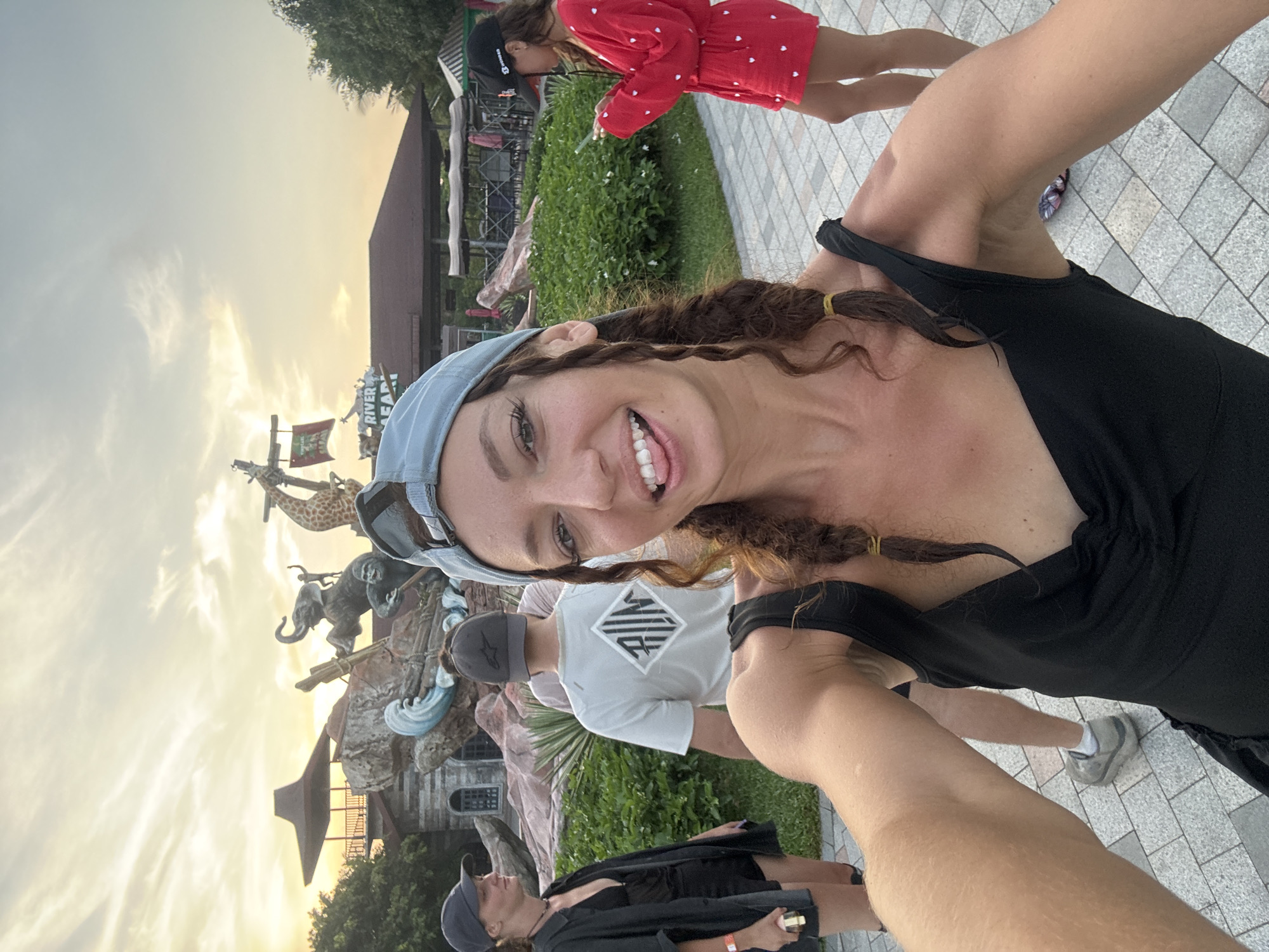 The image shows a selfie taken at the entrance of the River Safari Đà Nẵng in Vietnam. The woman in the foreground is smiling at the camera, wearing a black tank top and a blue baseball cap. Behind her are several people, including a child in a red dress with white hearts, a person in a white t-shirt with a logo, and another person wearing a black outfit and a hat. In the background, there is a large sculpture featuring various animals like a giraffe and an elephant, and a building that is part of the River Safari complex, all set against an early evening sky. The ground is paved with rectangular stones.