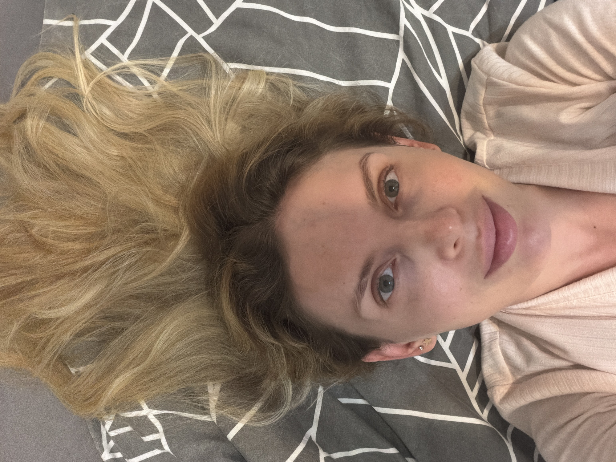 A woman is depicted in a close-up, high-angle portrait, appearing to be lying down and looking directly at the camera. Her long, wavy blonde hair is spread out behind her on a patterned surface. She has light-colored eyes, a fair complexion with scattered freckles, and a soft, relaxed expression featuring a slight smile with her lips gently parted. A small silver stud piercing is visible on her right nostril, and she wears small stud earrings on her visible ear. She is dressed in a light-colored, possibly cream or pale pink, long-sleeved top with a ribbed texture. The background is a gray fabric adorned with a white abstract geometric line pattern, suggesting a bedsheet or blanket. The lighting is soft and ambient, indicating an indoor setting. This image was taken in Budva, Montenegro.