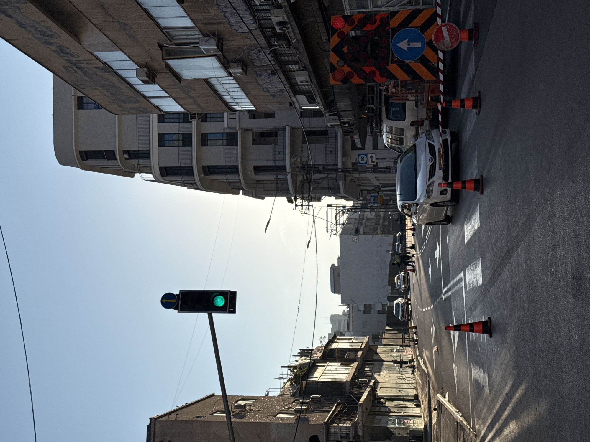 The image depicts a street scene in Tel Aviv, Israel, oriented with the sky at the lower left and the street extending into the upper right. A traffic light is visible, displaying a green light and a left-turn signal. Buildings line both sides of the street; the architecture includes both older structures and a taller, modern building. Traffic cones are placed along the right side of the road, near a white car and a construction sign with a blue left arrow and a red circle with a white line through it. Other vehicles can be seen further down the street. The asphalt of the road shows painted lane markers and areas of light and shadow. The sky is clear.