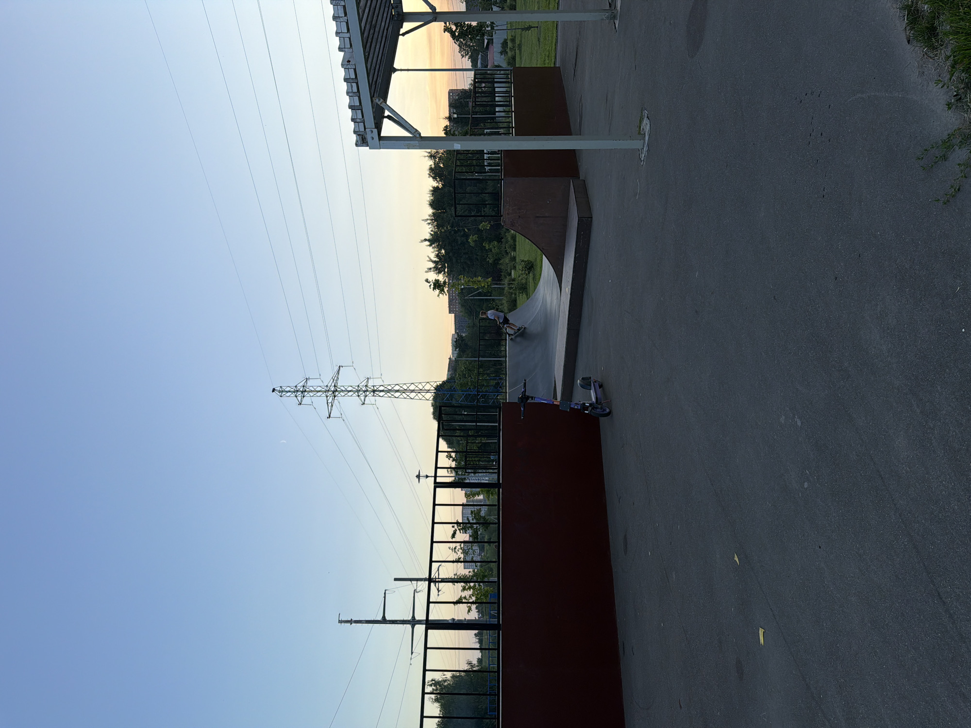 The image depicts a skate park in Shcherbinka, Russia, during what appears to be late afternoon or early evening. The sky is clear with a gradient of blue to a pale orange near the horizon. A prominent power line tower stands tall in the background, with several power lines stretching across the frame. In the foreground, a dark asphalt surface dominates, with a purple kick scooter parked on the left side of the image.
The skate park itself features a large, dark red ramp structure with a curved transition, made of wood and metal. A young person, wearing a white t-shirt and dark shorts, is seen riding a scooter down the ramp. To the right of the ramp, a covered structure with a metal frame and roof stands partially in view.
The environment surrounding the skate park includes trees and some distant buildings, suggesting an urban or suburban setting. A black metal fence encloses parts of the skate park area. The overall impression is one of a recreational space in use during a quiet time of day.