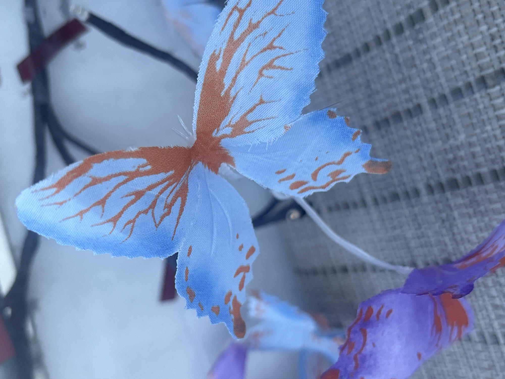 A close-up, shallow depth-of-field photograph captures a cluster of artificial butterflies, likely used as decoration or part of a costume. The prominent butterfly in the foreground has light blue wings with delicate, irregular burnt orange markings resembling veins or intricate patterns. The wing edges are frayed. Behind it, other butterflies are visible, including one with purple wings and similar orange markings, and another with light blue wings, slightly out of focus. A thin, white wire extends from the butterflies upwards, suggesting they are attached to a headband or a similar accessory. The background is blurred, showing dark wires and a red object, possibly indicating the presence of electronics or other decorative elements. The butterflies are resting on a textured surface that appears to be a grey mat or fabric, with a pattern of raised rectangular shapes. The lighting is soft, casting gentle shadows and highlighting the fabric texture of the butterfly wings. There is no indication of the specific location within Osterode am Harz, Germany.