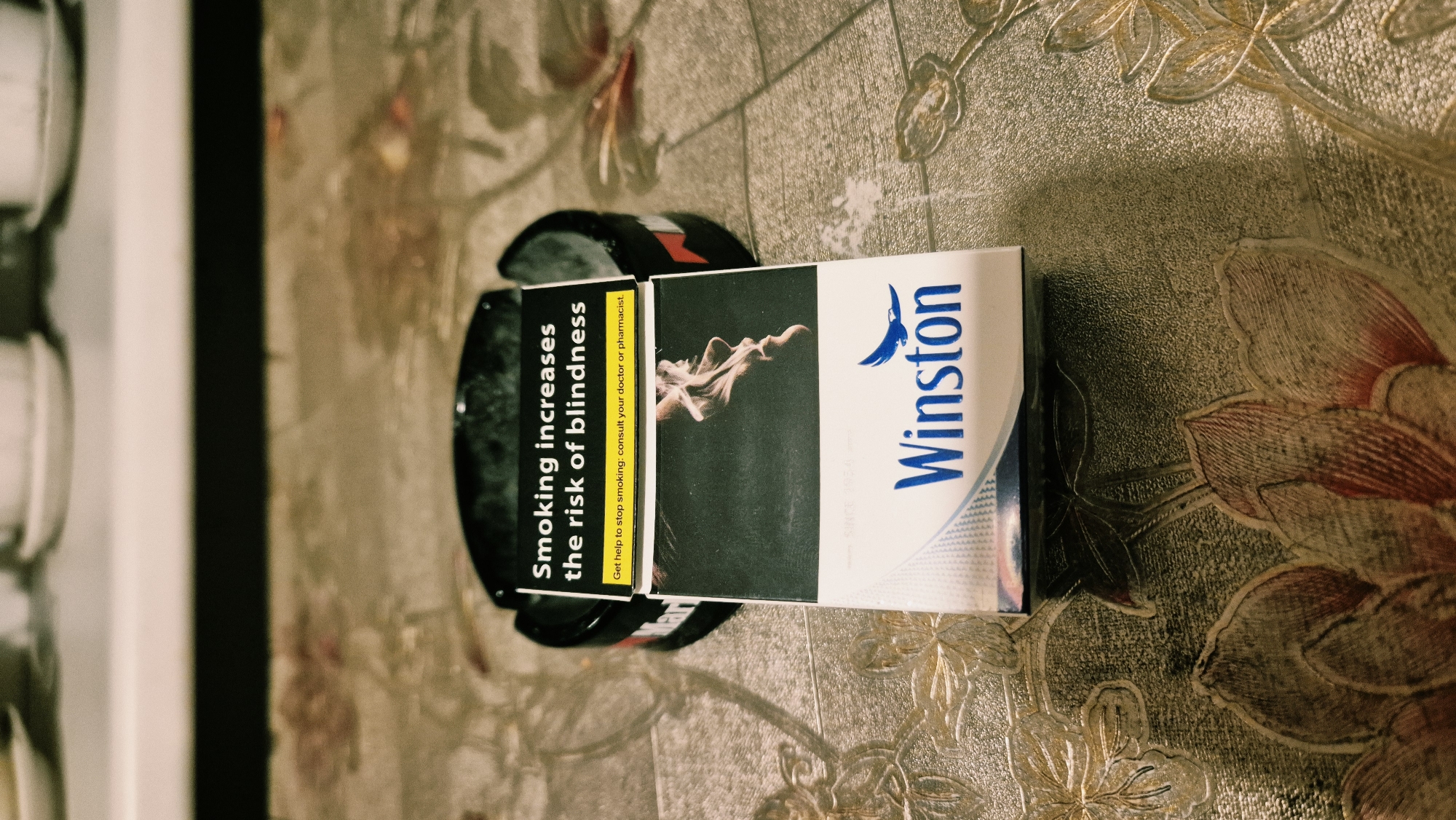 The image shows a pack of Winston cigarettes placed in an ashtray on a patterned tablecloth. The pack has a warning label that reads "Smoking increases the risk of blindness" and "Get help to stop smoking: consult your doctor or pharmacist." The background shows a blurred view of some cups and saucers on a shelf. The scene is indoors, on a table with a floral pattern. There are no people visible in the image. The lighting appears to be artificial. The location of Varena, Lithuania is not evident from the image itself.
