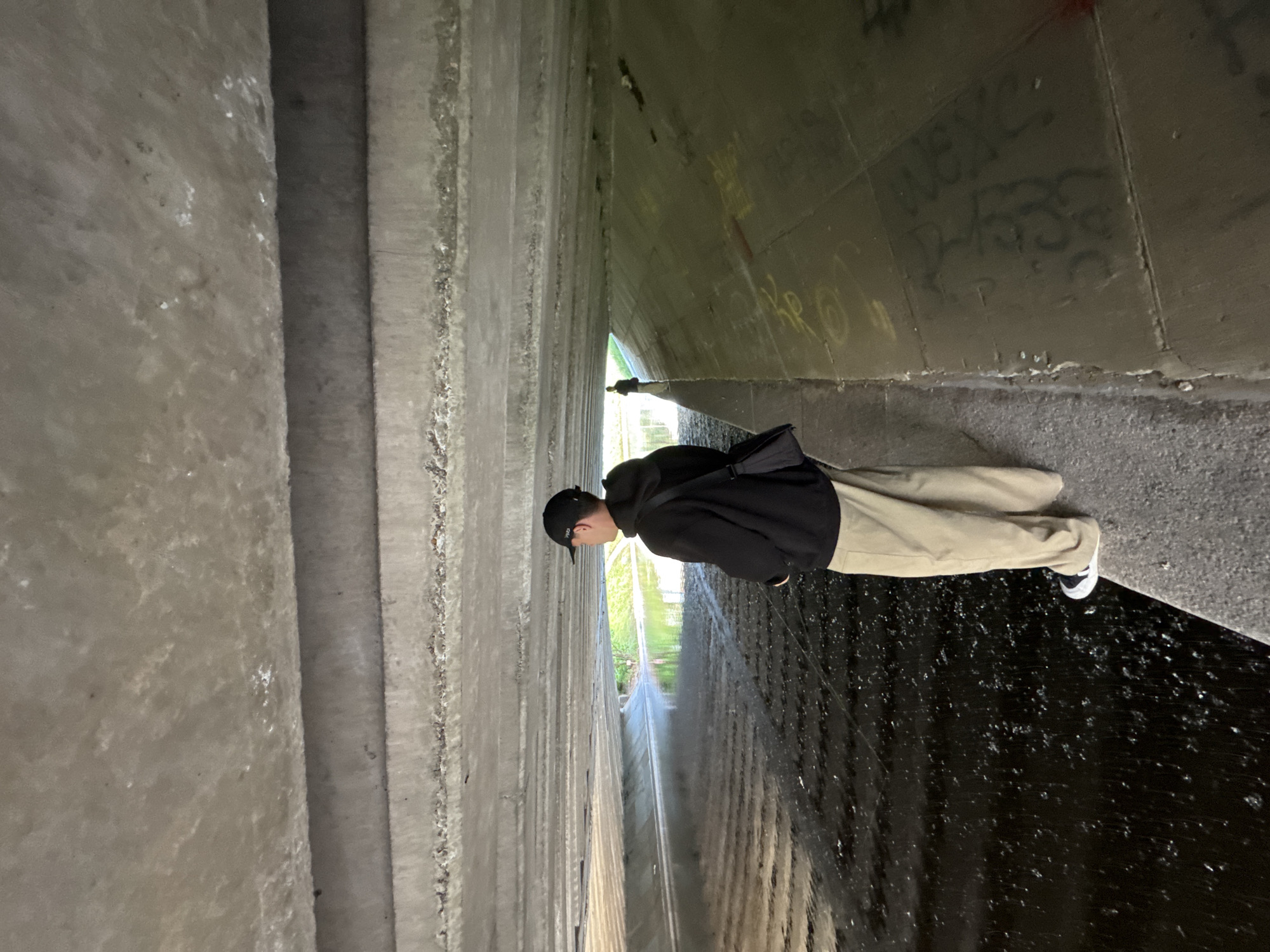 An adult male stands facing away from the camera, looking out at a water channel under a concrete overpass. He is wearing a black hoodie, tan pants, a black baseball cap, and a black crossbody bag. His left foot is on the edge of the concrete embankment. The water in the channel is dark and rippled, with reflections of green foliage visible in the distance. The concrete structure above is made of multiple horizontal beams, and the side walls of the channel have graffiti on them. Another person is visible in the distance further down the channel. The scene appears to be an urban drainage canal.