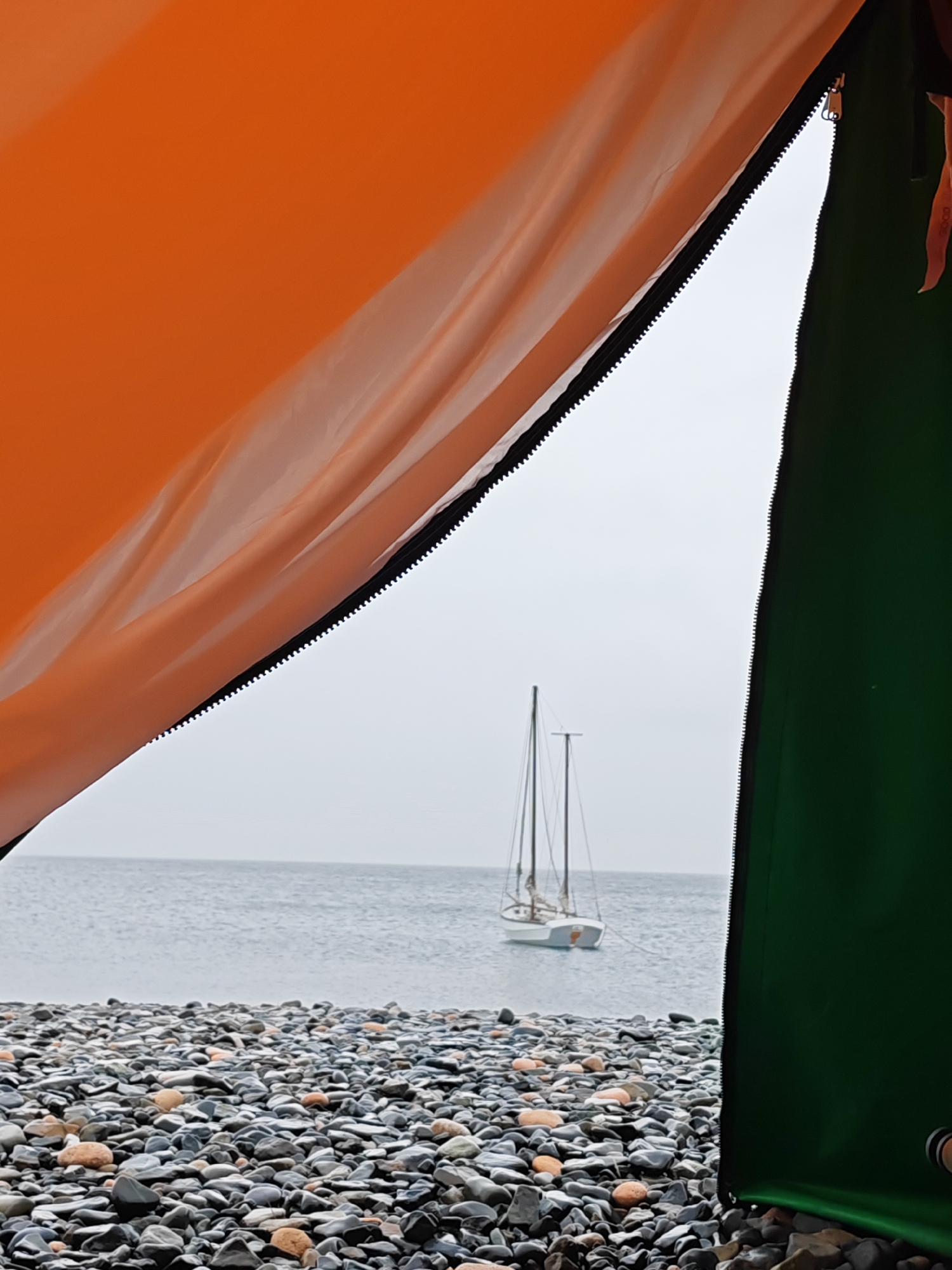 The image depicts a view through an open tent flap, looking out towards the ocean. The tent fabric is an orange color on the left and green on the right, with a zipper visible along the seam. In the distance, a small sailboat with two masts is anchored on calm, grey water under an overcast sky. The foreground is a rocky beach composed of smooth, grey and orange stones. The scene is devoid of human presence. The location is not specified as being in Reineke, Russia.