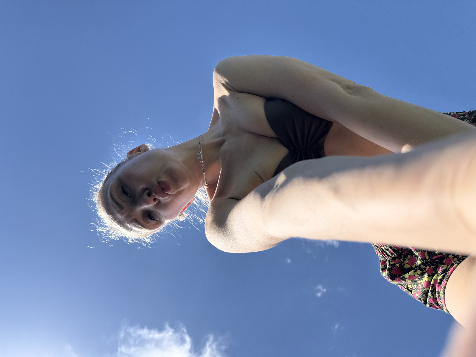 A low-angle shot shows a person with fair skin, blonde hair, and light eyes, wearing a dark bikini top and floral-patterned bottoms. The person is looking down at the camera with pursed lips and a possible pouting expression. Their arms are positioned in front of their chest. Behind them, a clear blue sky with a few wispy clouds is visible. The photograph gives the impression of being taken outdoors in bright daylight. There is no evidence in the image to confirm the location is Nizhnyaya Chermoda, Russia.