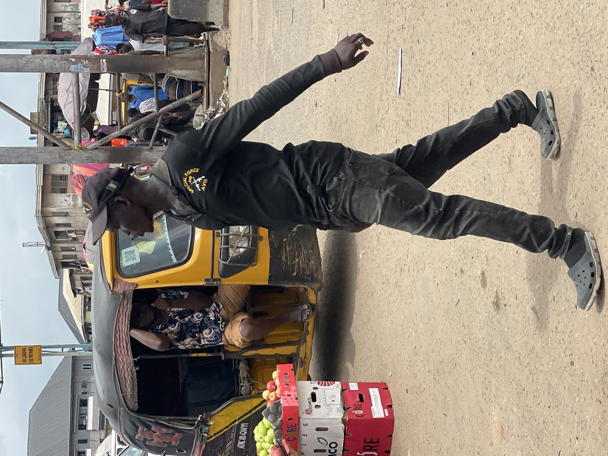 The image shows a man in black clothing, including a long-sleeved shirt with a "Special Force" logo and a baseball cap, standing beside a yellow tricycle in Onitsha, Nigeria. The tricycle is parked on a street with stacked boxes of apples in front of it. Another person is seated inside the tricycle. A sign that reads "NO LOADING USE THE PARKS" is visible. Other people and buildings are in the background, creating a busy urban street scene.