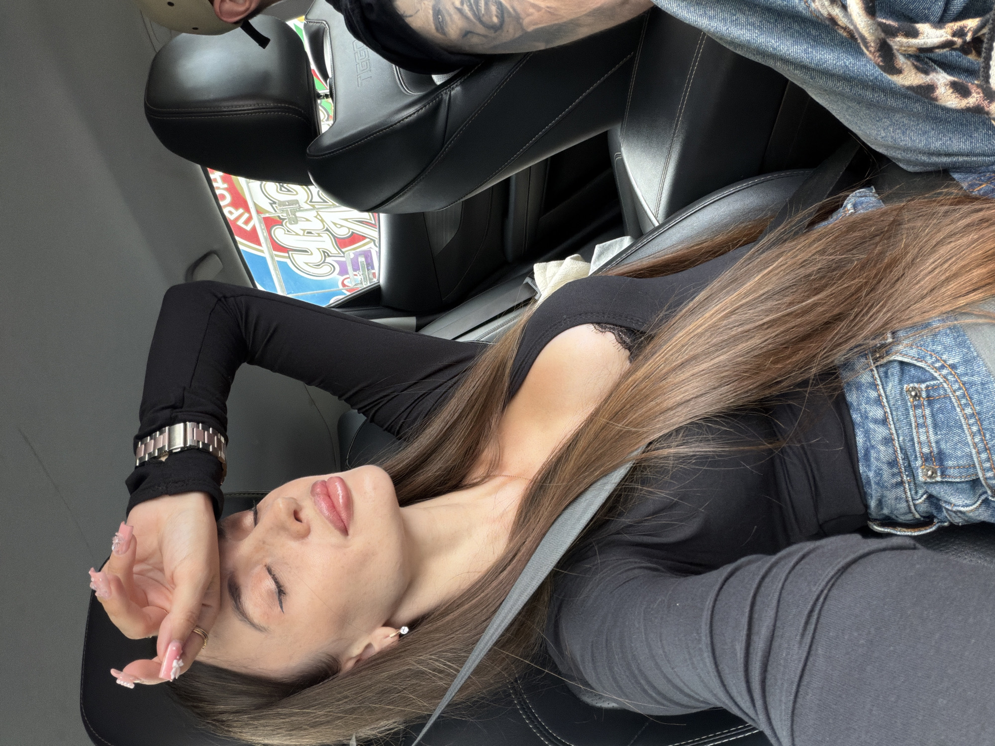 The image shows a young woman with long brown hair sitting in the passenger seat of a car. She is wearing a black long-sleeved top and blue jeans. Her eyes are closed, and her right hand is resting on her forehead. A seatbelt is across her chest. The interior of the car, including black leather seats, is visible. Outside the car window, a colorful sign with Russian text is partially visible. Another person is in the driver's seat, with part of their arm and a tattoo visible. The background is blurred, but appears to be a daytime outdoor scene.