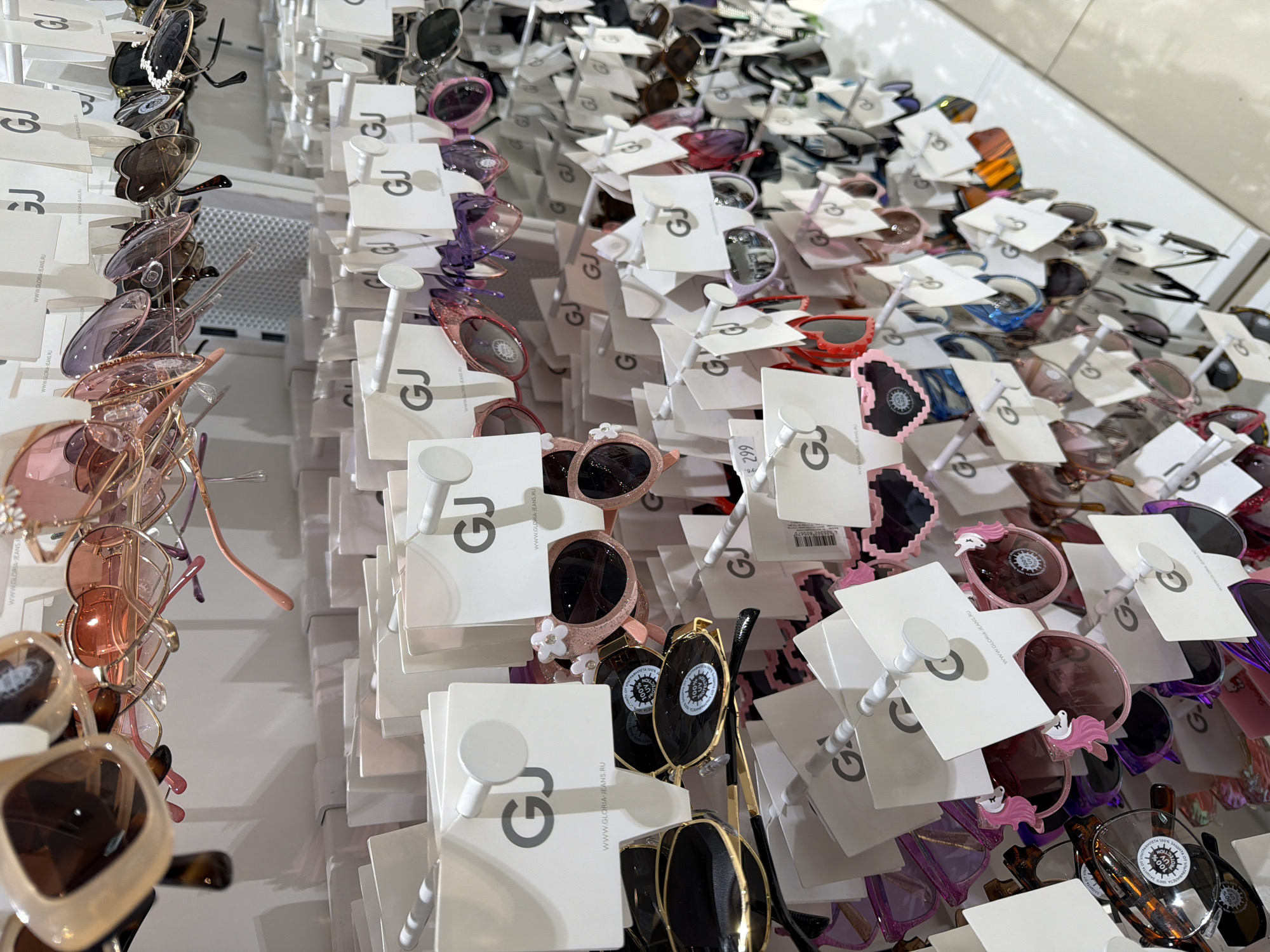 This image shows a display of sunglasses for sale. The sunglasses are arranged on a wall, each pair hanging from a white card with the logo "GJ" on it. The sunglasses come in a variety of styles and colors, including black, pink, purple, and tortoiseshell. The environment appears to be a retail store. Based on the OCR, the logo suggests that the location is a Gloria Jeans store in Chaikovsky, Russia.