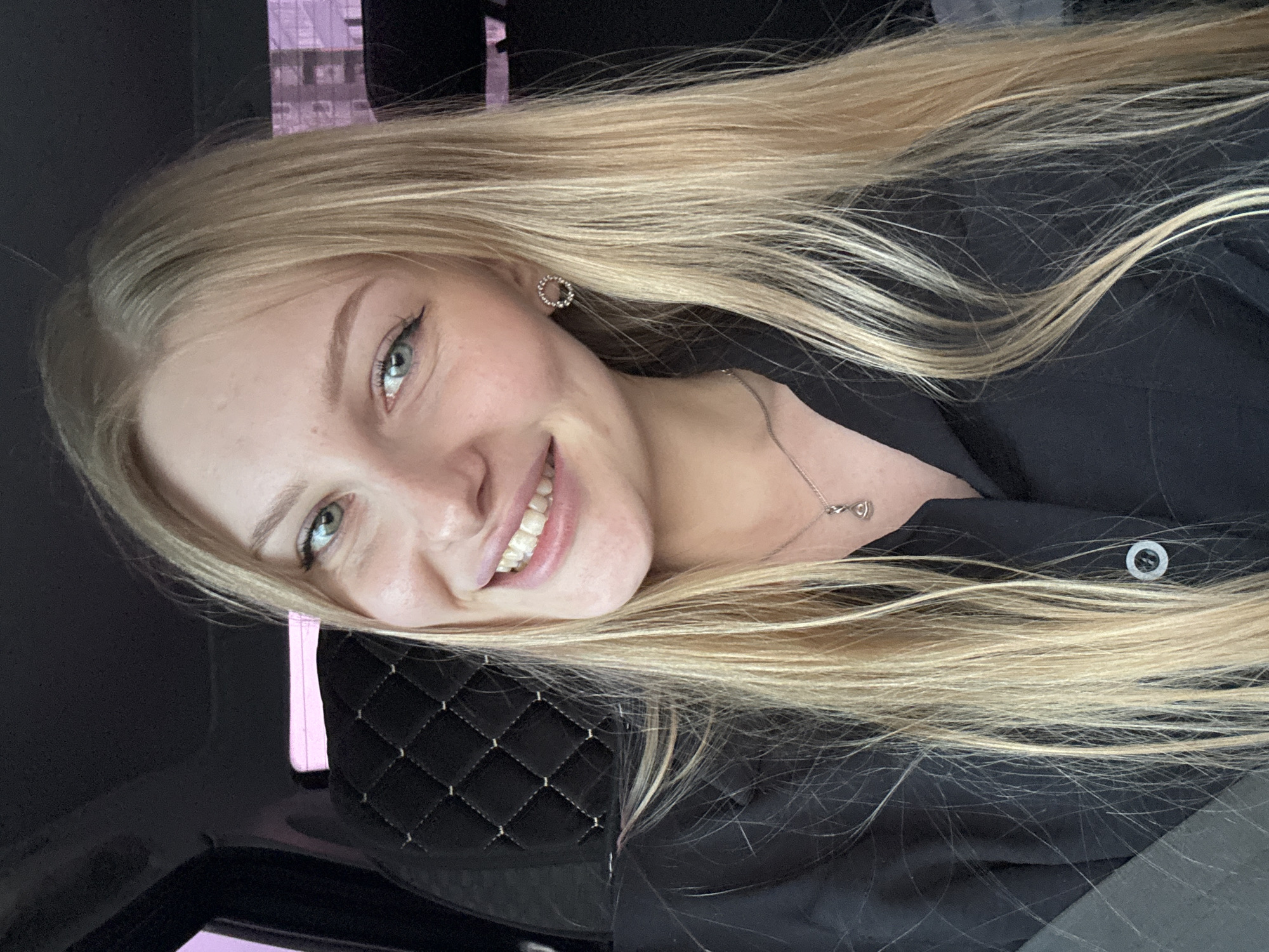 A light-skinned woman with long blonde hair is smiling. She is wearing a black shirt or jacket and a silver necklace. She also wears a silver earring. The background is blurry and dark, suggesting she is indoors, possibly in a car with quilted seat. The location is Кондратово, Russia.