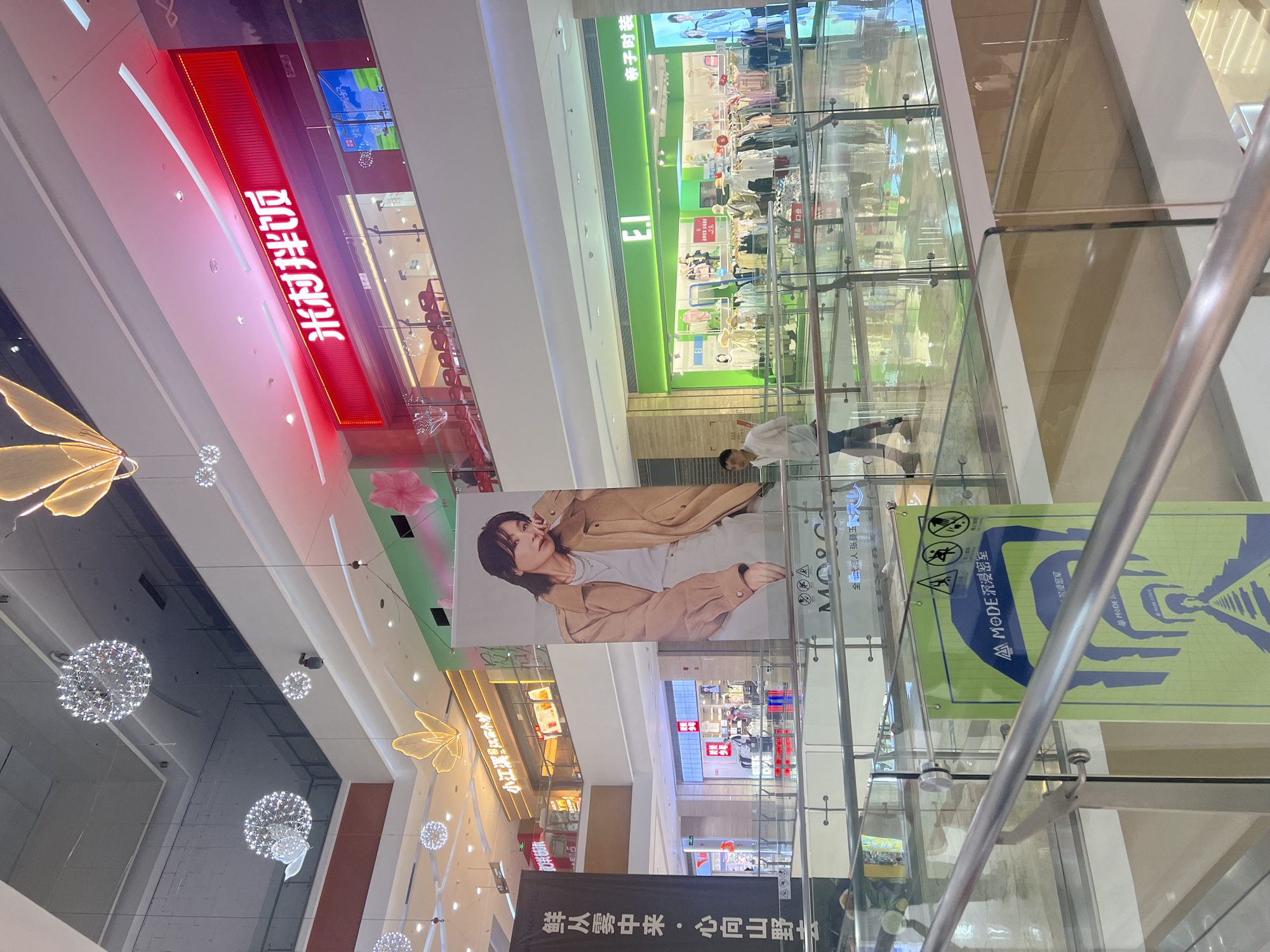 The image depicts the interior of a modern shopping mall, likely in Liandu District, China. The perspective is from an upper level walkway, looking across to various storefronts and a large banner featuring a woman.
In the foreground, a glass railing runs along the walkway, reflecting the light and the surrounding environment. A portion of a green banner with blue graphics and text in Chinese is visible on the lower left, advertising "MODE Immersion Room."
Across the mall, several shops are visible. A prominent green storefront displays the logo "E.I." and "亲子服装" (children's clothing). Racks of clothing are displayed both inside and outside the store. Further back, another store with a red illuminated sign that reads "米村拌饭" (Micun Banfan, a Korean restaurant) is situated on a higher level. Smaller shops with various displays are also visible in the background.
A large banner hanging in the center of the frame features a portrait of a woman wearing a trench coat. Above the shops, decorative elements are suspended from the ceiling, including spherical light fixtures resembling chandeliers and illuminated butterfly-shaped decorations.
A man in a white shirt and dark pants is walking across a glass floor on the right side of the frame. The mall's architecture includes multiple levels with walkways and open spaces, creating a sense of depth and scale. The lighting is bright and diffuse, characteristic of an indoor shopping environment.