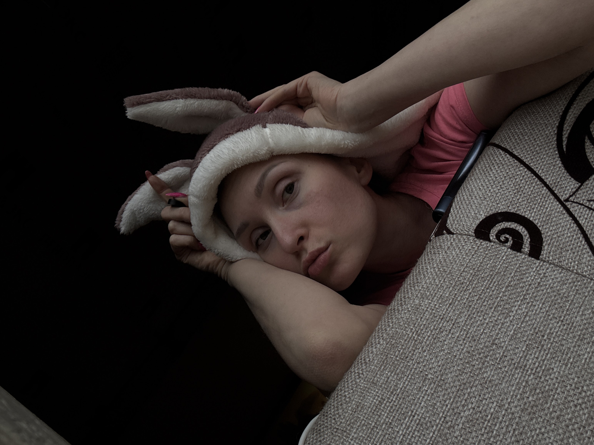 A person is pictured wearing a brown and white rabbit-ear hat, resting on a gray patterned couch with a dark background. The person has a pink shirt on, and their hands are near their head. A dark, rectangular object is slightly visible between the person and the couch. The photograph appears to be taken at an angle. The location Nizhnyaya Chermoda, Russia cannot be verified from the provided image.