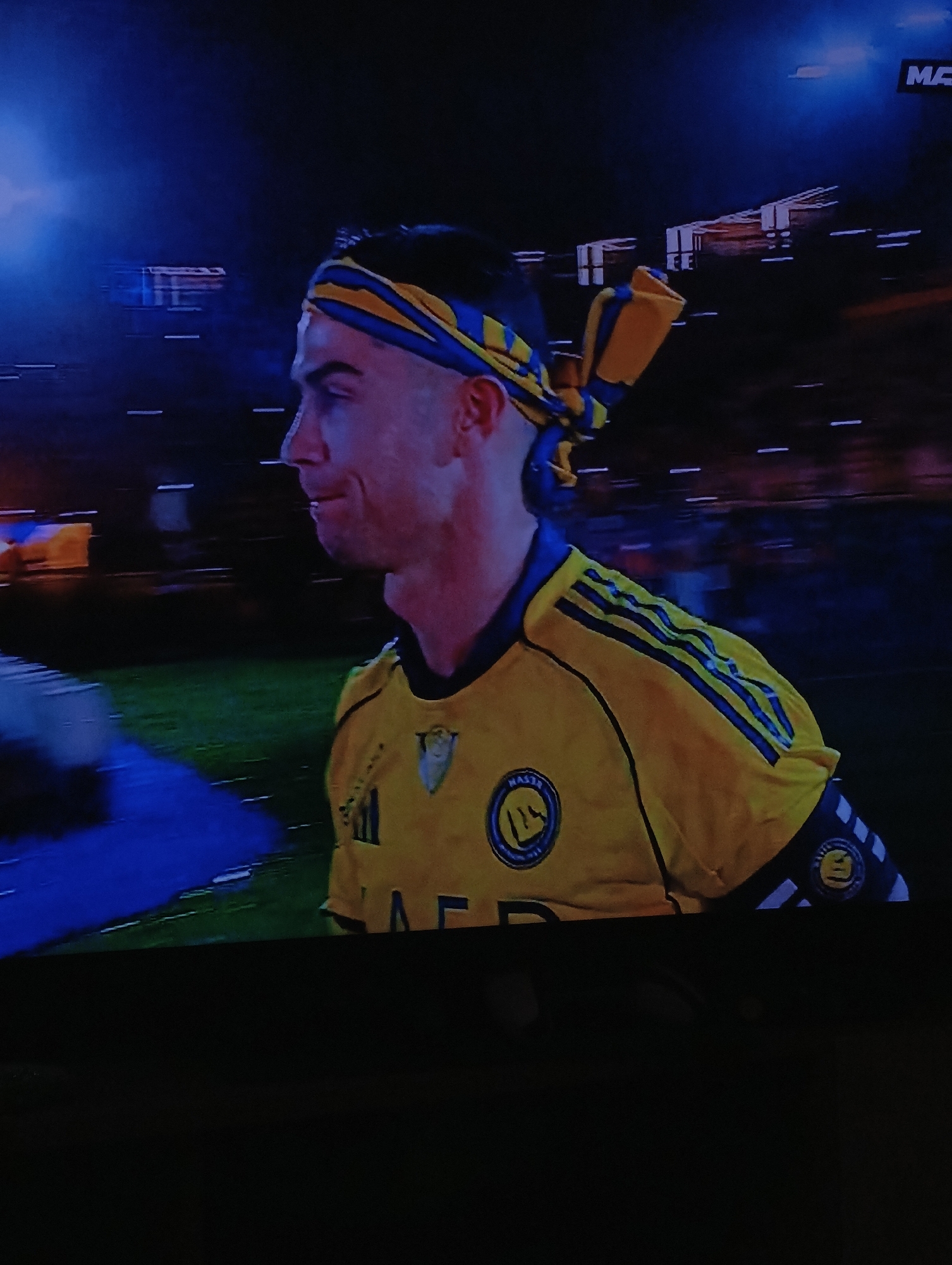 The image shows a soccer player wearing a yellow jersey with blue stripes on the sleeves and shoulders, and a blue and yellow headband. The jersey has the word "NAEE" printed on it. The soccer player is standing on a field, and the background appears to be a stadium with blurred lights and spectators. The location in the city of Plovdiv, Bulgaria is not specified. The player seems to be in motion.
