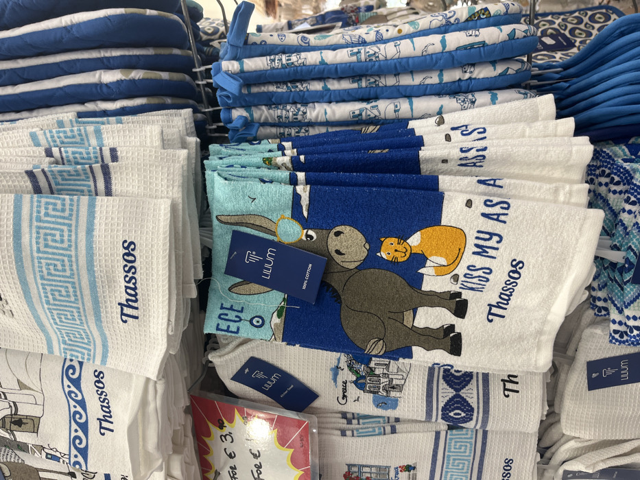 The image displays stacks of souvenirs, seemingly for sale. On the left, there's a stack of white waffle-textured towels with blue Greek key designs and the text "Thassos". Beside them, another stack features towels with cartoonish designs of a donkey and a cat, along with the text "KISS MY AS A, Thassos." Some towels have tags that say “LILIUM, 100% COTTON". The bottom of the frame shows a price tag for "€ 3.90". In the background, there are stacks of blue and white patterned pot holders.