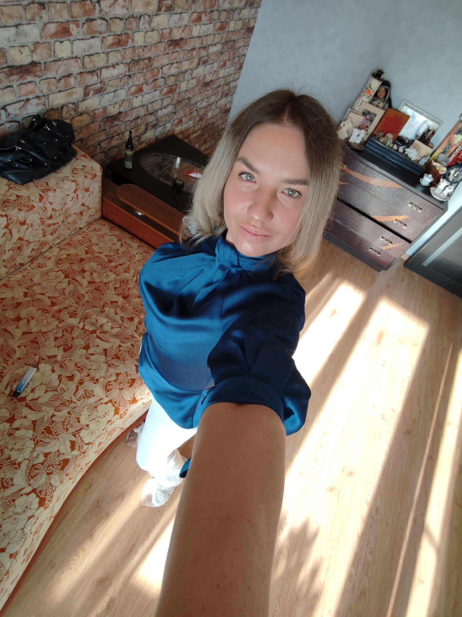 The image captures a woman taking a selfie indoors, likely in Petropavlovsk-Kamchatsky, Russia. The woman, with shoulder-length blonde hair and striking light green eyes, is looking directly at the camera with a calm expression. She is wearing a dark teal, high-necked blouse and white trousers. Her arm, extended to hold the camera, is visible in the foreground.
The setting is an indoor room, likely a living area or bedroom. To the left, an ornate orange and cream floral patterned sofa is visible, with a black handbag adorned with studs resting on it. Next to the sofa is a dark wooden end table with a glass top, holding a dark bottle and a red item. A brick-patterned wallpaper covers the wall behind the sofa. To the right, a dark wooden dresser laden with framed photos, figurines, and other decorative items is visible. The wall behind the dresser is a plain light gray.
The room features a light wooden floor. Strong diagonal streaks of sunlight and window frame shadows fall across the wooden floor, indicating a bright morning or afternoon. A small, light-colored object, possibly a remote, rests on the patterned sofa cushion near her feet. No discernible text is visible in the image.