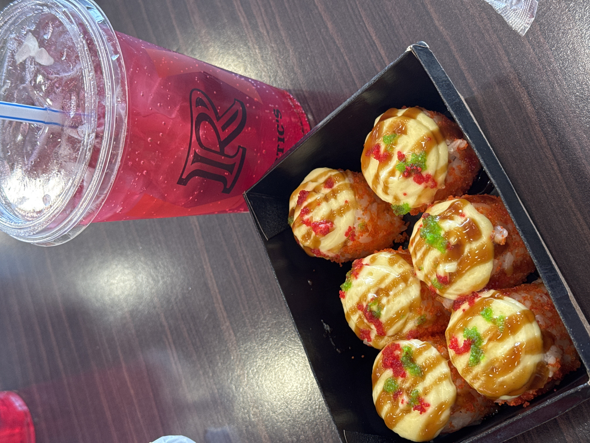 An overhead shot captures a beverage and a box of food on a dark brown, wood-grained table, likely in a casual dining setting. The scene is illuminated by bright, diffused lighting, suggesting daytime indoors.
On the left, a clear plastic cup with a lid and a blue-and-white striped straw holds a vibrant red, fizzy drink with ice cubes. A black, stylized "R" logo is prominent on the side of the cup, with the letters "TIC'S" visible below it, suggesting a brand name like "ARCTIC'S" or similar.
To the right, a black rectangular box contains several pieces of food. These appear to be fried sushi rolls or similar rice-based bites, individually topped with a generous dollop of a light yellow, creamy sauce, a zigzag drizzle of a darker brown sauce (possibly teriyaki or unagi sauce), and colorful sprinkles. The sprinkles are a mix of bright red and green, resembling roe or decorative sugar. The base of the food items, partially visible, seems to be rice coated in red or orange fish roe (tobiko/masago).
The objects are neatly arranged, indicating they are ready to be consumed. No people are visible, and no specific emotional context can be inferred. The image portrays a food and drink combination at what is contextually identified as a location in Moscow, Russia.