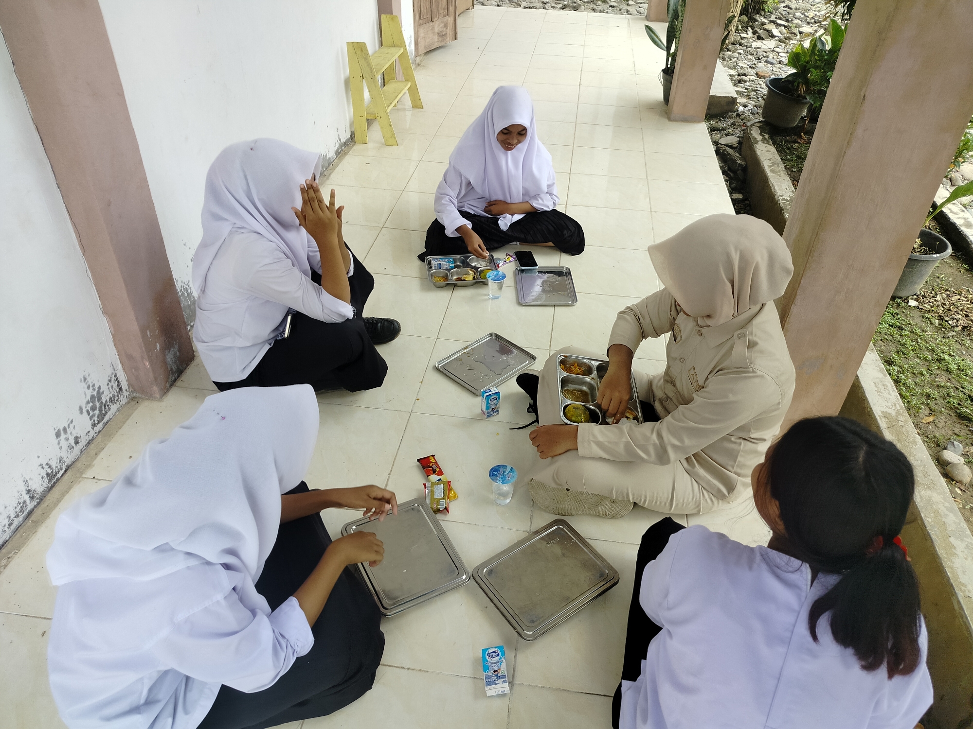Four young females, all wearing white hijabs and school uniforms, are sitting on a tiled outdoor walkway. They are gathered around metal trays containing food. One girl in the center, facing the camera, is smiling and appears to be eating. To her left, another girl is covering her face with her hands. To her right, a girl in a tan uniform is also eating from her tray. The fourth girl, facing away from the camera, is wearing a white uniform and has a ponytail. The scene takes place outside a building with a pink and white exterior wall and wooden doors. Plants in pots are visible to the right, and a simple wooden shelf unit stands against the wall. The overall setting suggests a school or educational institution in Purwodadi, Indonesia.