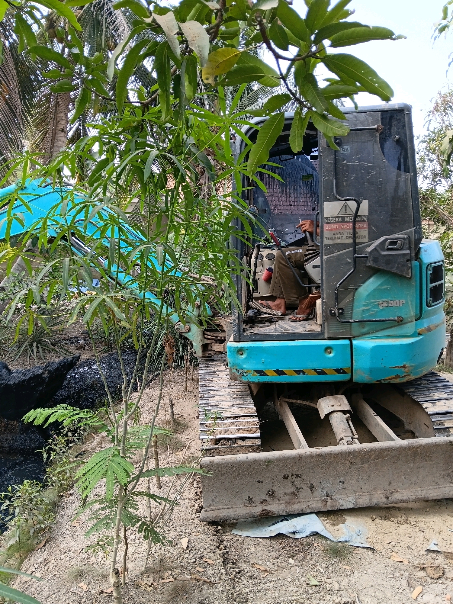 An outdoor scene in Margo Rukun, Indonesia depicts a blue excavator, partially obscured by foliage. A person wearing brown pants and flip-flops is seated in the excavator's cab, holding a cigarette. A sign on the cab reads "SEGERA MENU... BLIND SPOT AREA". The excavator's tracks and front blade are visible, resting on a dirt and gravel surface. To the left of the excavator, a black object, possibly a chicken, is situated near dark, wet ground. Palm trees and other vegetation frame the scene, suggesting a tropical or subtropical environment.