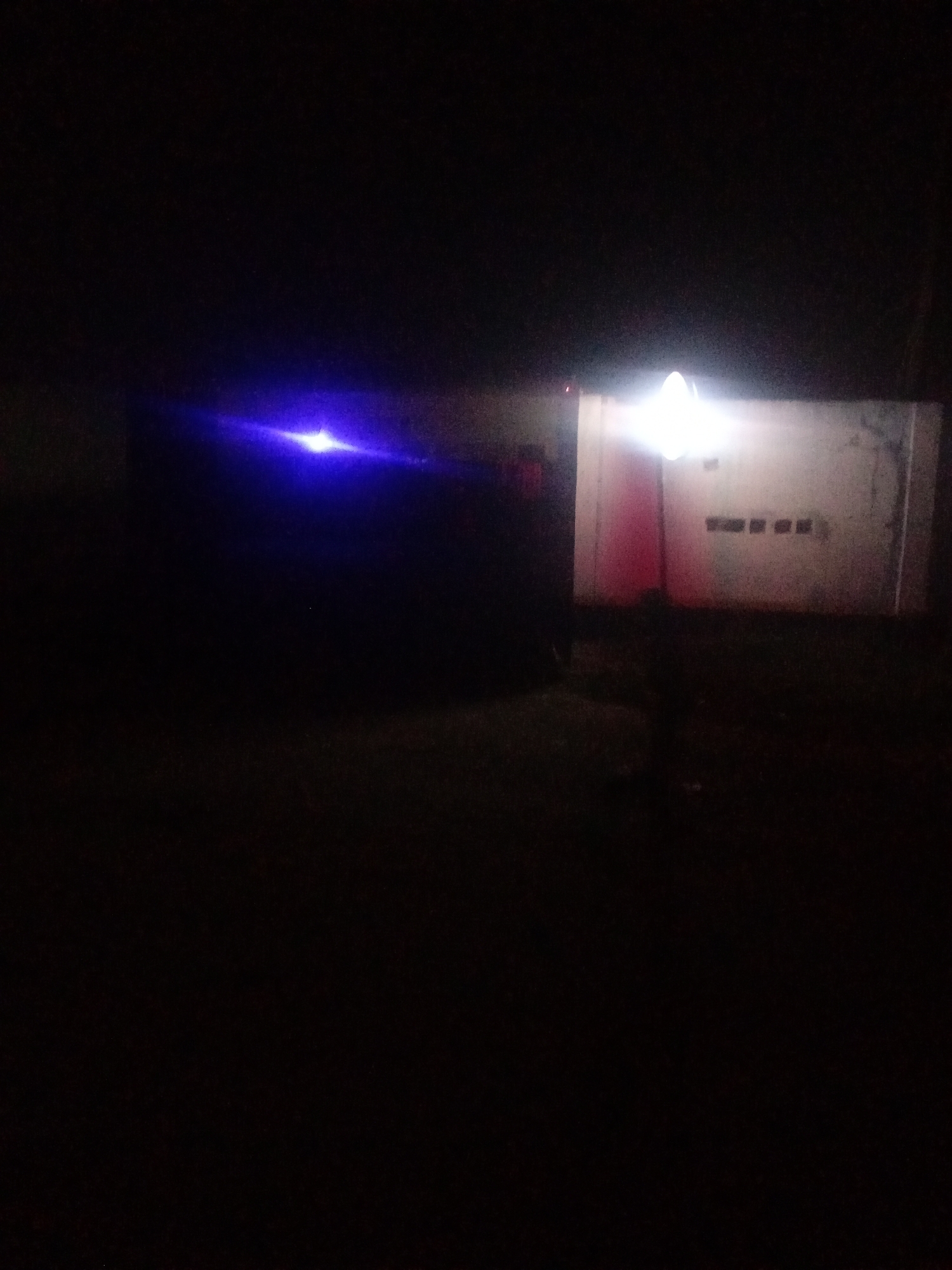 The image depicts a dimly lit outdoor scene at night, possibly in the city of Sabon Gari, Nassarawa, Nigeria. A bright, possibly blue, light source is visible in the upper left quadrant, casting a strong glare. To the right, a building with a light-colored wall is illuminated by a bright white light emanating from above. The wall has several dark, rectangular shapes that could be windows or openings. A reddish vertical element, potentially a door or painted section, is visible on the left side of the building. The foreground and surrounding ground area are dark, with minimal detail visible due to the low light conditions. The overall impression is one of a poorly lit, possibly industrial or commercial area at night.
