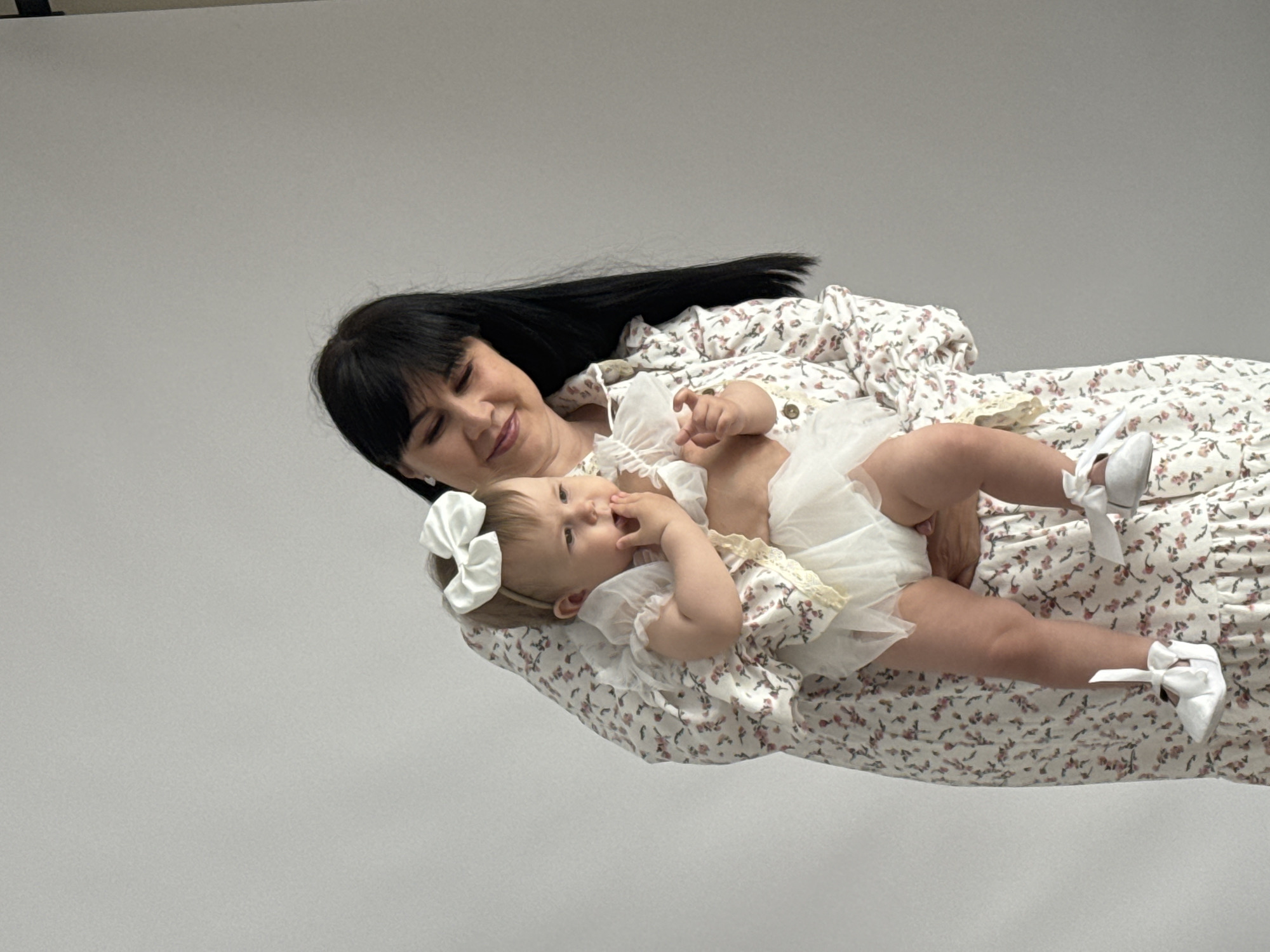 A medium shot shows a woman with black hair wearing a floral dress, lying sideways on a white surface with a baby on her chest. The baby is wearing a white dress with lace trim and a white bow headband, and white shoes with bows. The woman is looking down at the baby with a gentle smile. The background is a plain white wall. The image was not taken in Izhevsk, Russia.
