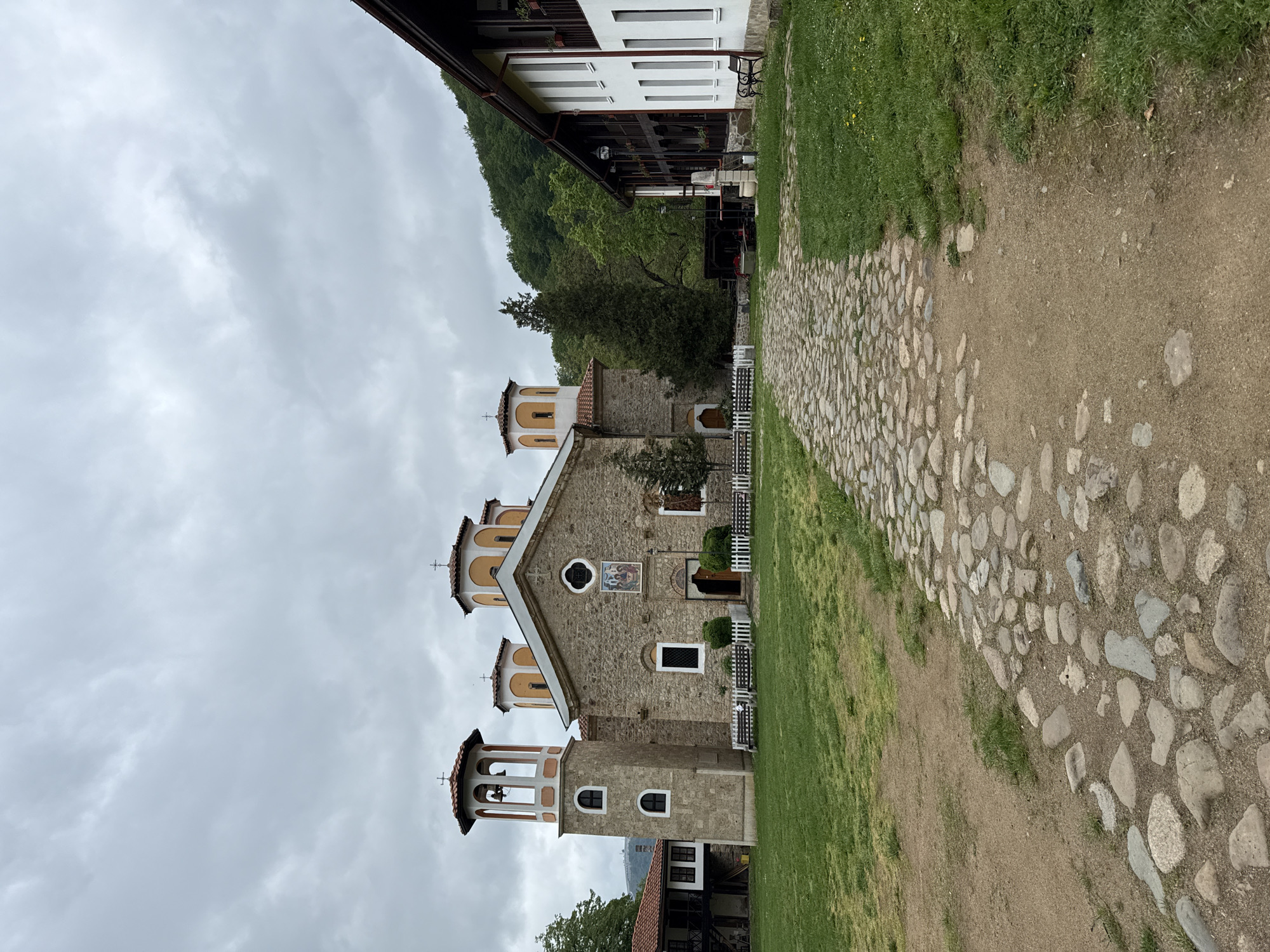 The image shows a stone church located in Etropole, Bulgaria. The church has three towers, each topped with a cross. The towers are painted in beige and white. The church building is stone. A white picket fence surrounds the church. A stone path runs alongside the church. The sky is overcast. A building with white siding is in the background. A grassy hill is also visible in the background.