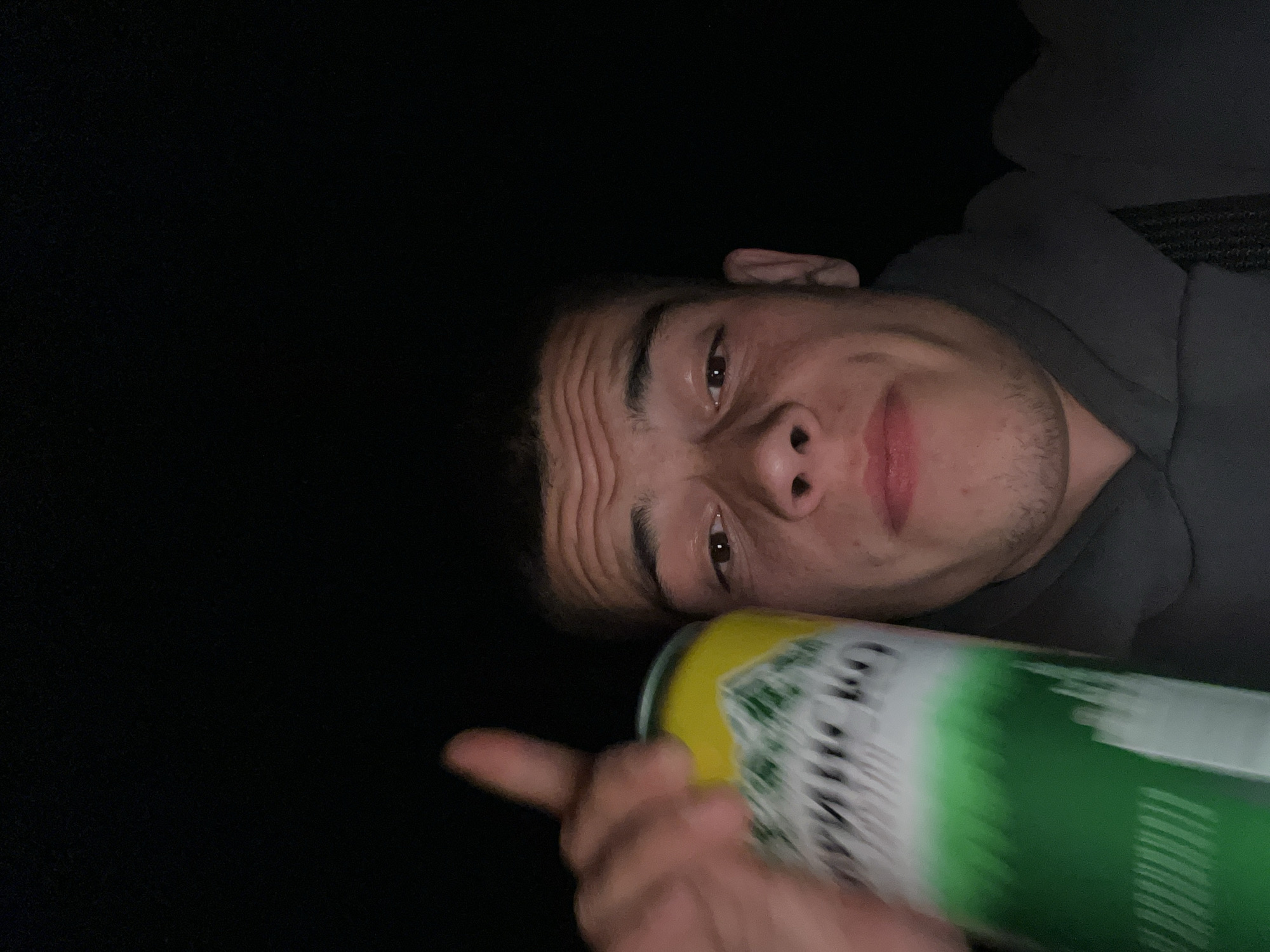 A man is shown close-up, partially reclined and angled towards the camera. His face has visible wrinkles on his forehead. He is wearing a dark-colored hooded sweatshirt. His right hand is holding a green and yellow can, pointed at the camera. The background is dark. It is not possible to determine if this image was taken in the city of Karavelovo, Bulgaria from the visual information provided.