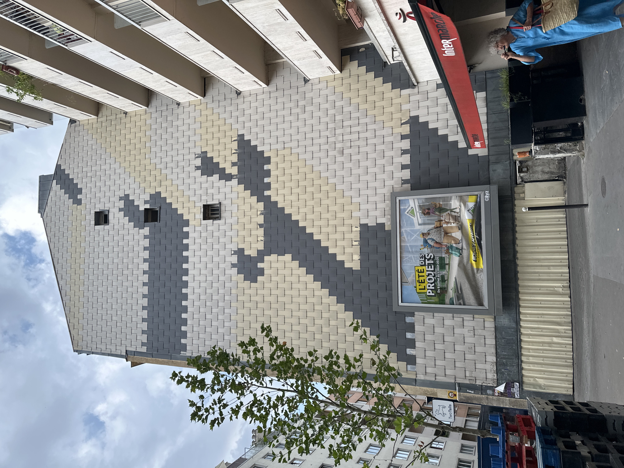 The image shows a street scene in Paris, France, with a large building facade covered in a geometric tile pattern of gray, white, and beige. A billboard advertising "L'ÉTÉ DES PROJETS" is visible at the bottom of the building. To the right, a red awning with the "Intermarché" logo extends from a store. A woman in a blue dress and carrying a straw bag is standing on the sidewalk, adjusting her glasses. A tree with green leaves is on the left side of the image, partially obscuring the building. In the foreground, there are stacks of blue and red plastic pallets. The sky is overcast with some clouds.