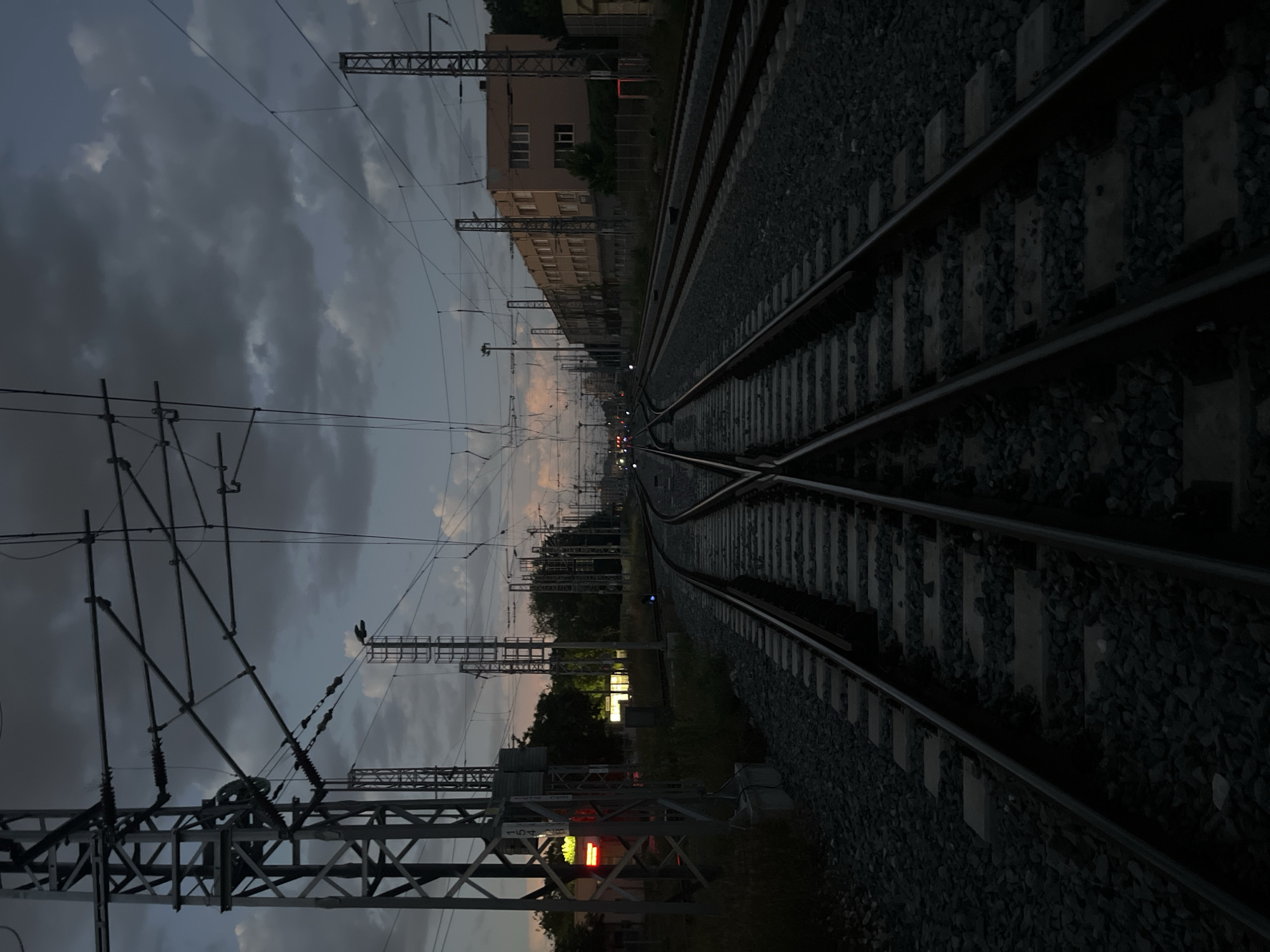 The image depicts a railway track scene in an urban environment. The tracks converge in the distance, flanked by ballast stones. Overhead power lines and support structures are visible against a backdrop of a cloudy sky. A multi-story building stands parallel to the tracks on one side. Signal lights are present along the track. Vegetation appears in the mid-ground.