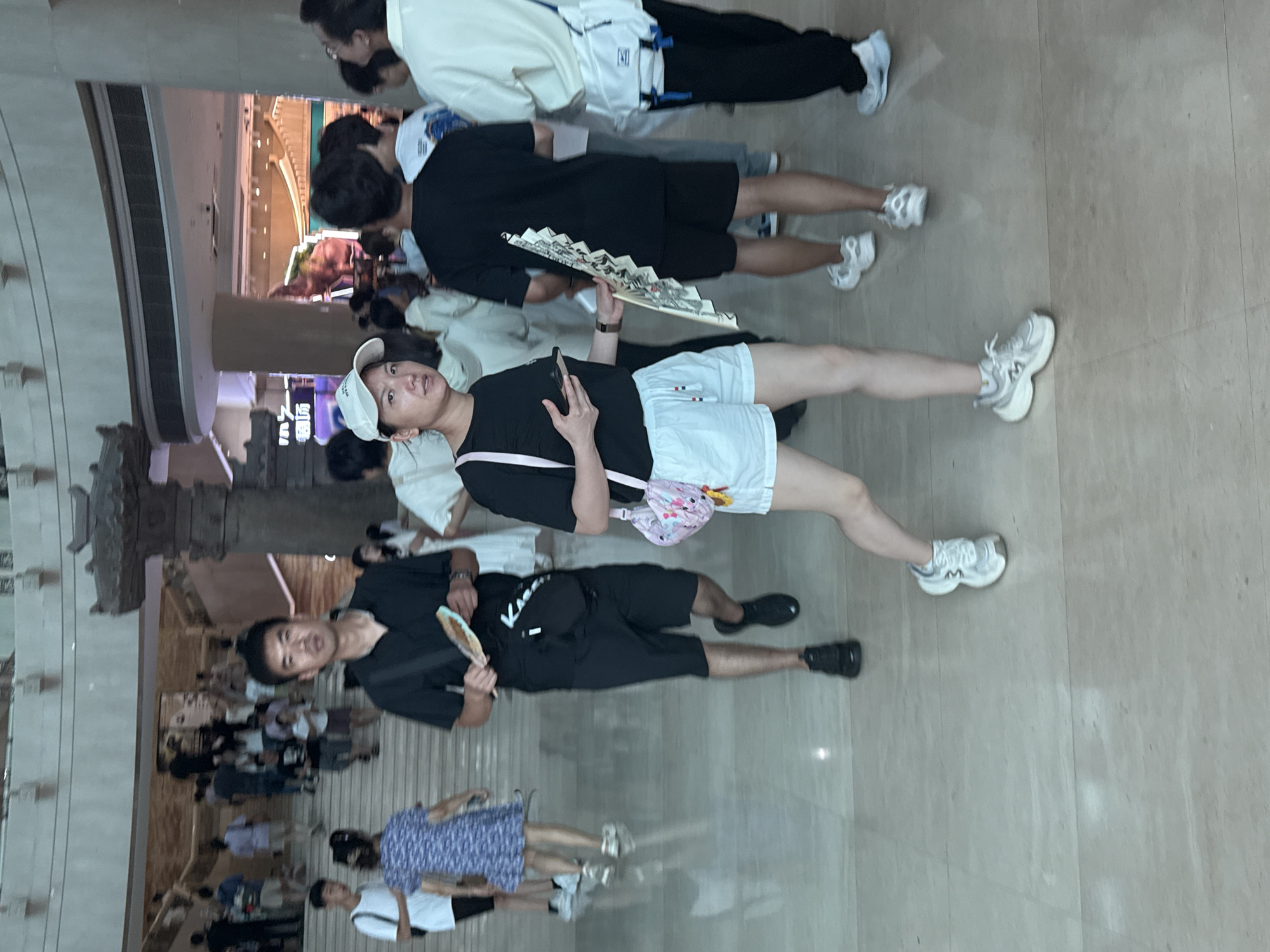 In Yuzhong District, China, a group of people are gathered inside a large, multi-level building, likely a museum or cultural center, judging by the architectural style and large ornamental pillar visible. The floor is made of polished light-colored stone. In the foreground, a woman wearing a white cap, black t-shirt, and white shorts walks with a pink purse on her left shoulder and a smartphone in her right hand. To her left, a man in a black polo shirt and black shorts walks with a fan in his right hand and a black crossbody bag. Behind them, other individuals are seen walking up and down escalators and along the hallways, with some carrying fans as well. The atmosphere appears casual, with many people dressed in summer attire.