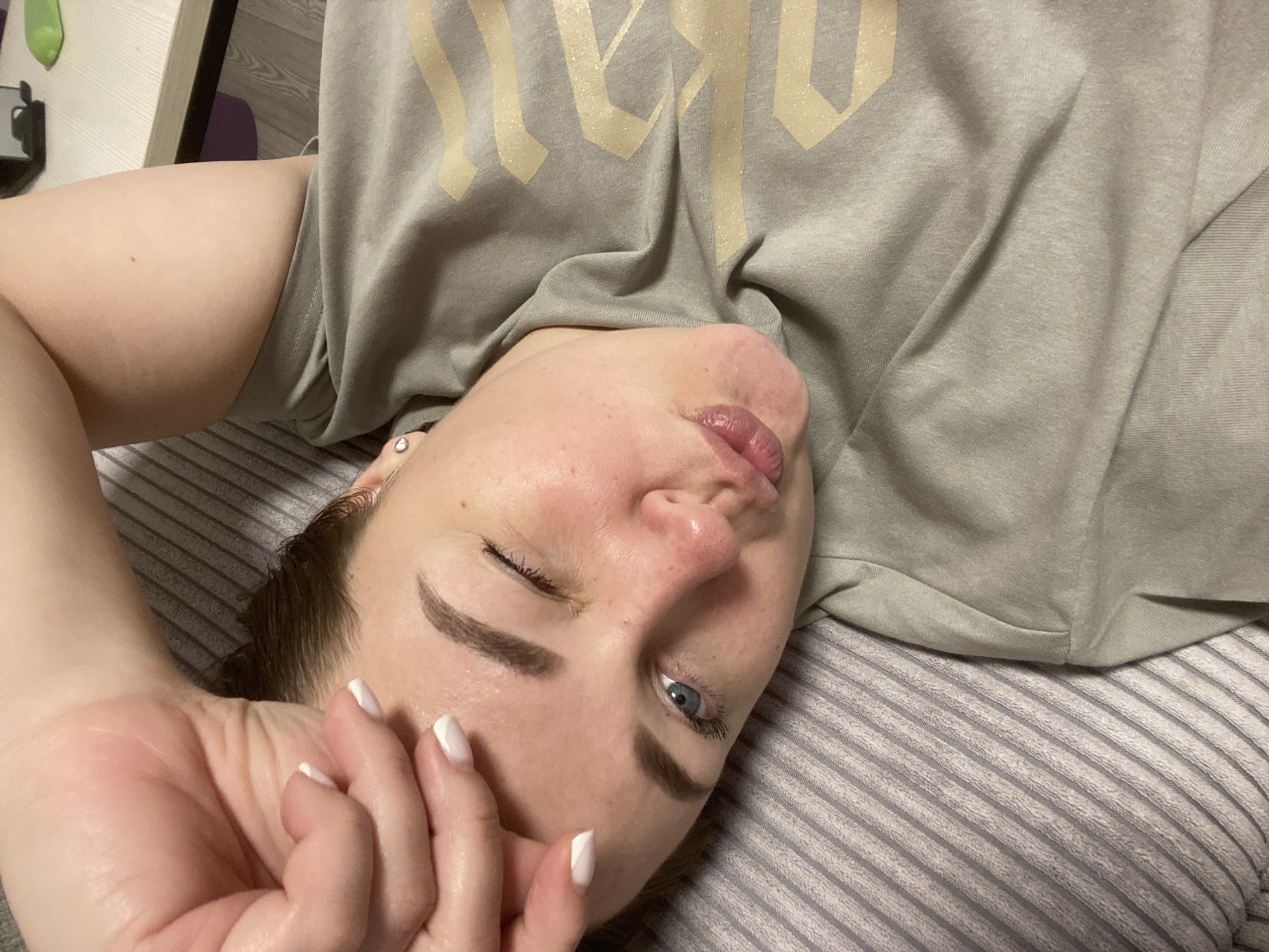 A person, likely female, is captured lying down indoors, possibly on a bed or sofa, in a relaxed pose. The image is taken from a slightly elevated, diagonal perspective. Her face, positioned towards the top right, features light skin, prominent dark eyebrows, and one open blue eye with mascara, while the other eye appears closed or winking. Her lips are slightly parted and puckered.
Her left hand, adorned with white-tipped fingernails and a ring featuring a small clear stone, rests near her face. A small silver earring is visible on her left earlobe. She is wearing an oversized, light greyish-green short-sleeved t-shirt. The t-shirt has a large, stylized text design in a light gold or glittery yellow color, which appears to spell "WARRIOR" across the chest. A portion of her bare upper thigh is visible below the t-shirt, suggesting she might be wearing shorts or underwear.
The background consists of striped bedding in grey and white, and parts of a dark wooden surface, possibly furniture like a table or headboard, along with a lighter surface. The lighting indicates an indoor setting. The overall mood conveyed is relaxed and perhaps playful. The provided location context for this image is Sochi, Russia, though no explicit visual cues within the image confirm this.