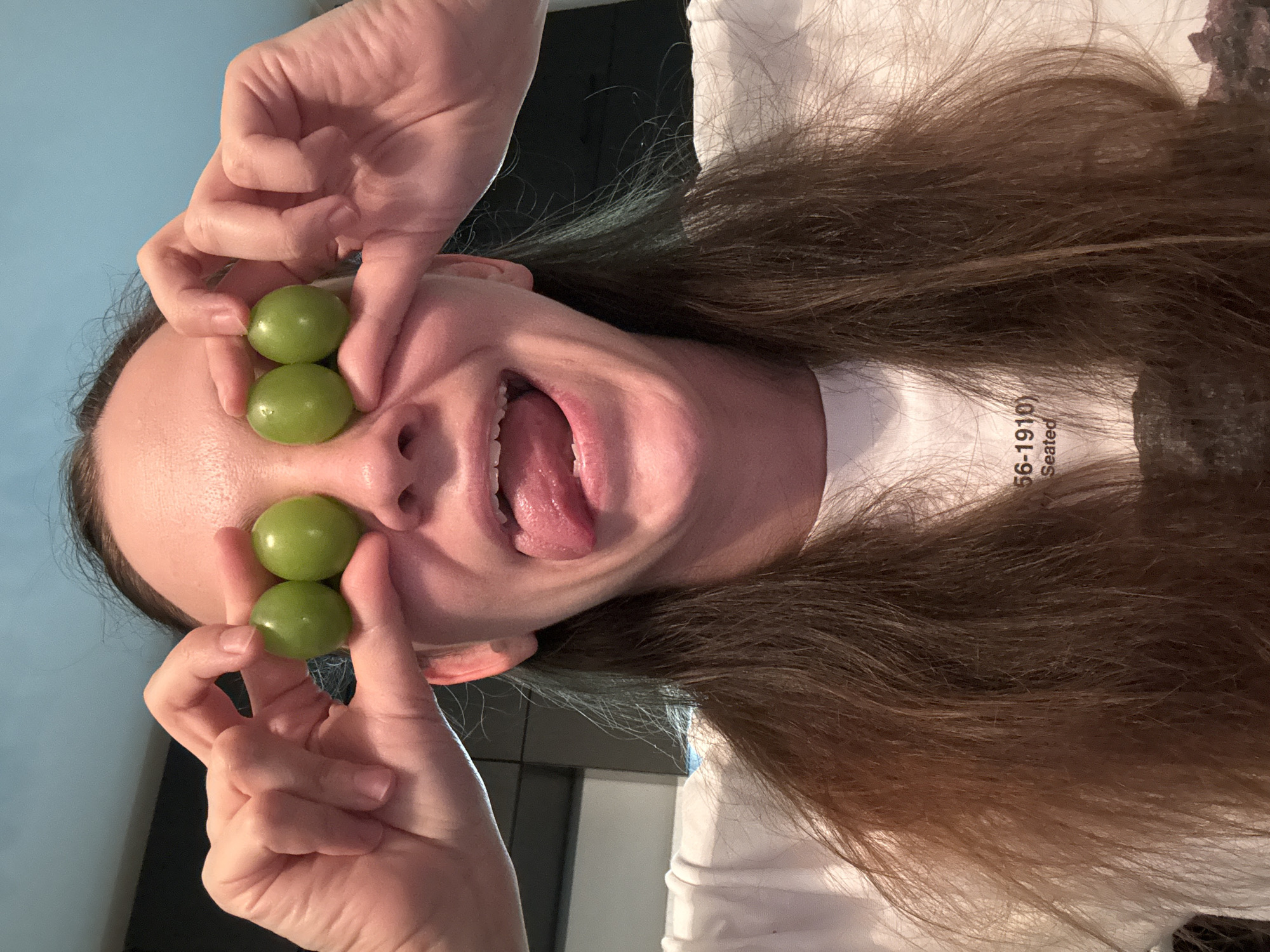A young woman with long brown hair is holding two green grapes over each of her eyes, making a playful expression with her tongue sticking out. She is wearing a white t-shirt with some text on it. The background appears to be an indoor setting with a blue wall and dark cabinetry. No location in Chaikovsky, Russia is visible.